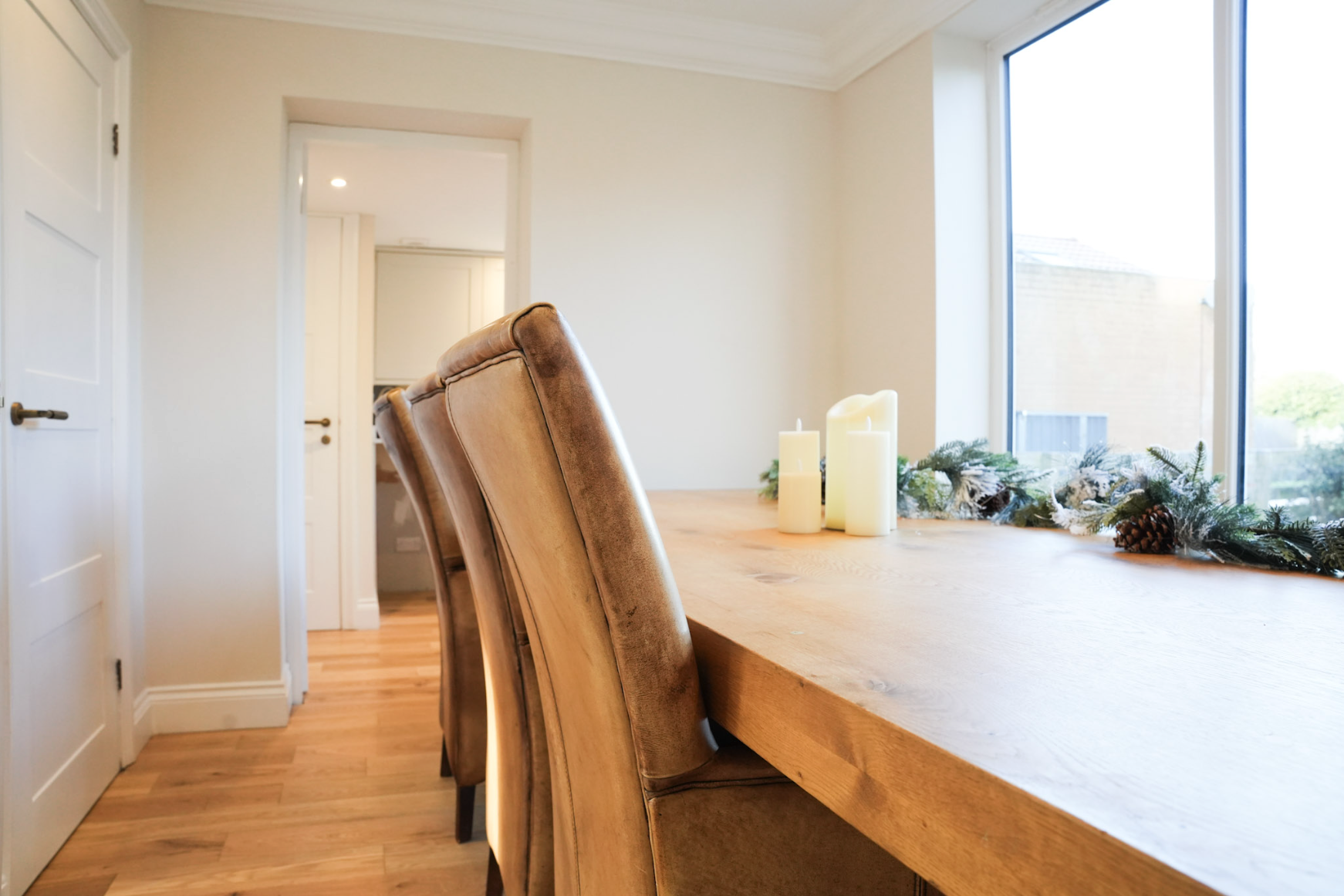 A wooden dining table decorated with candles and greenery, with leather chairs and a large window in a bright, modern dining room.