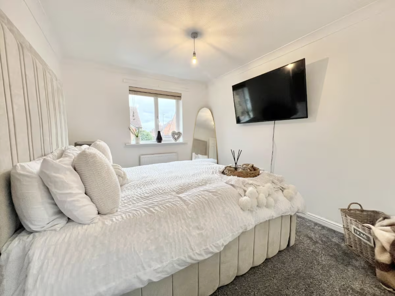 A cozy bedroom with a large bed covered in white bedding and pillows, a window with a view of rooftops, a mirror, and a flat-screen TV mounted on the wall.
