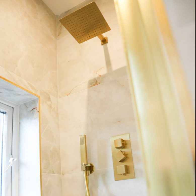 Gold-colored shower head and controls in a bathroom shower with beige marble tiles.