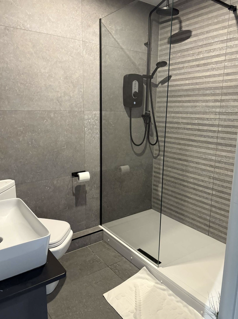 Bathroom with a walk-in shower, gray tiled walls, a white toilet, and a square white sink on a black countertop. There is a white bath mat on the gray tiled floor.