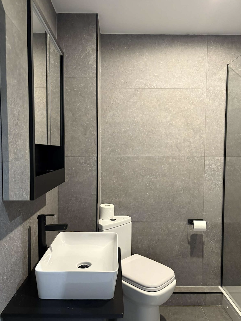 A modern bathroom with gray tiled walls, a white square vessel sink with a black faucet, a black countertop, a toilet with a roll of toilet paper on top, a wall-mounted mirror cabinet, and a toilet paper holder on the wall.