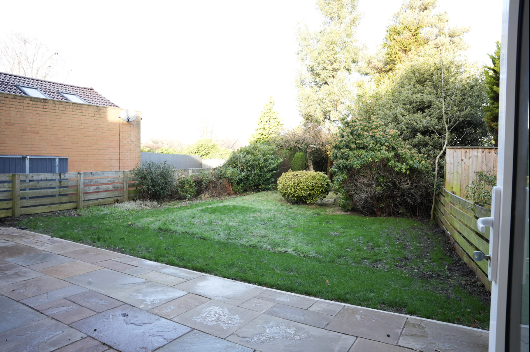 View of a backyard garden with green grass, bushes, trees, and a wooden fence, seen from an open patio with stone tiles.