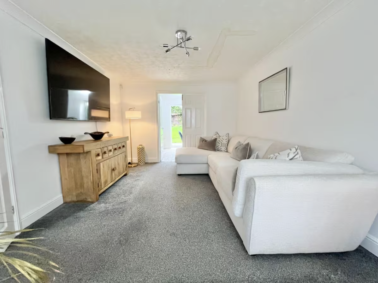 Living room with white sectional sofa, gray and patterned pillows, flat screen TV on wooden console, floor lamp, directional ceiling light fixture, door open to backyard, framed mirror on wall, gray carpeted floor, and white walls.