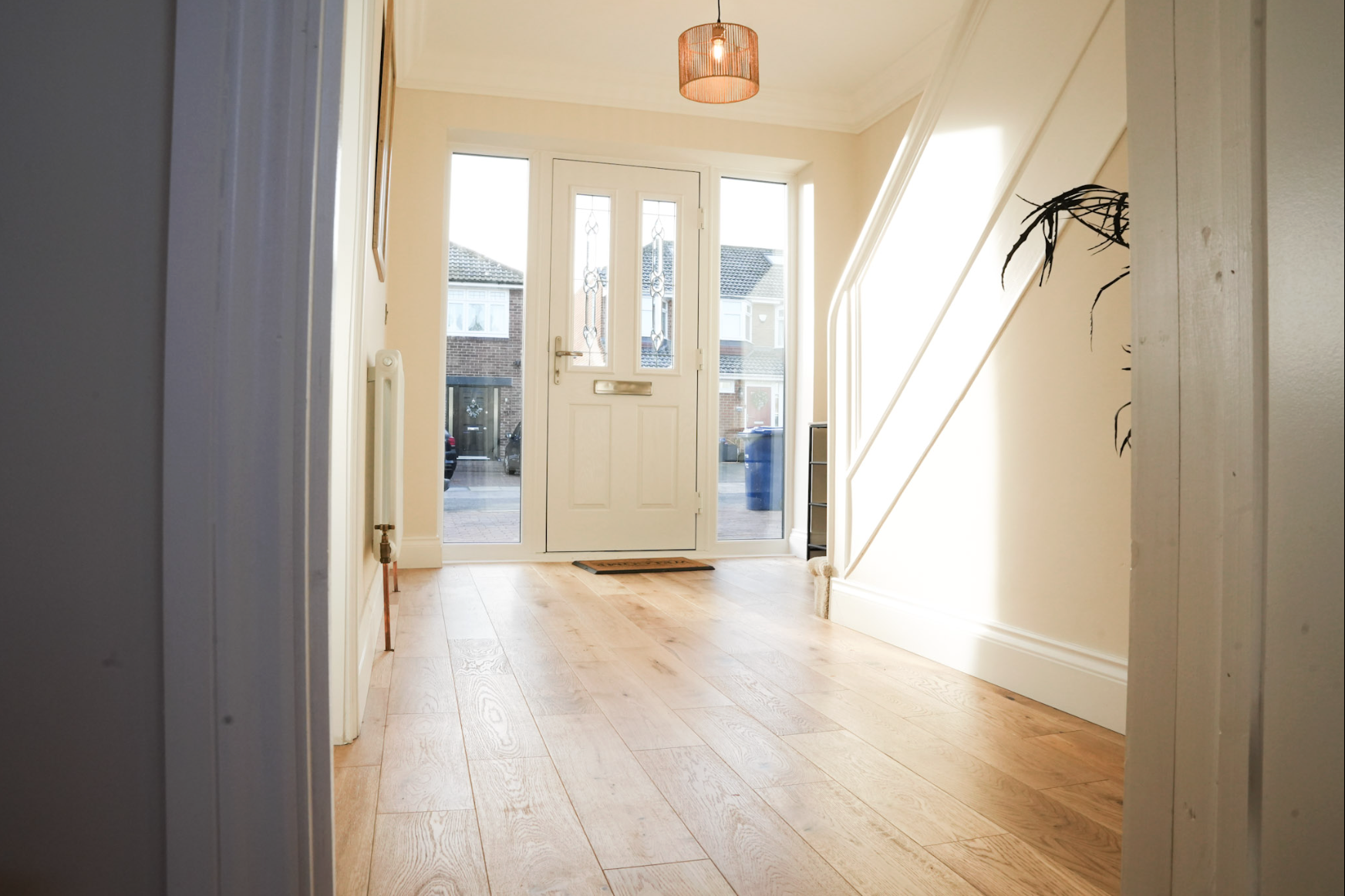Bright entrance hall with wooden flooring, glass-paneled front door, and a small black shelf on the right side. There is a ceiling light fixture and a radiator on the left side.