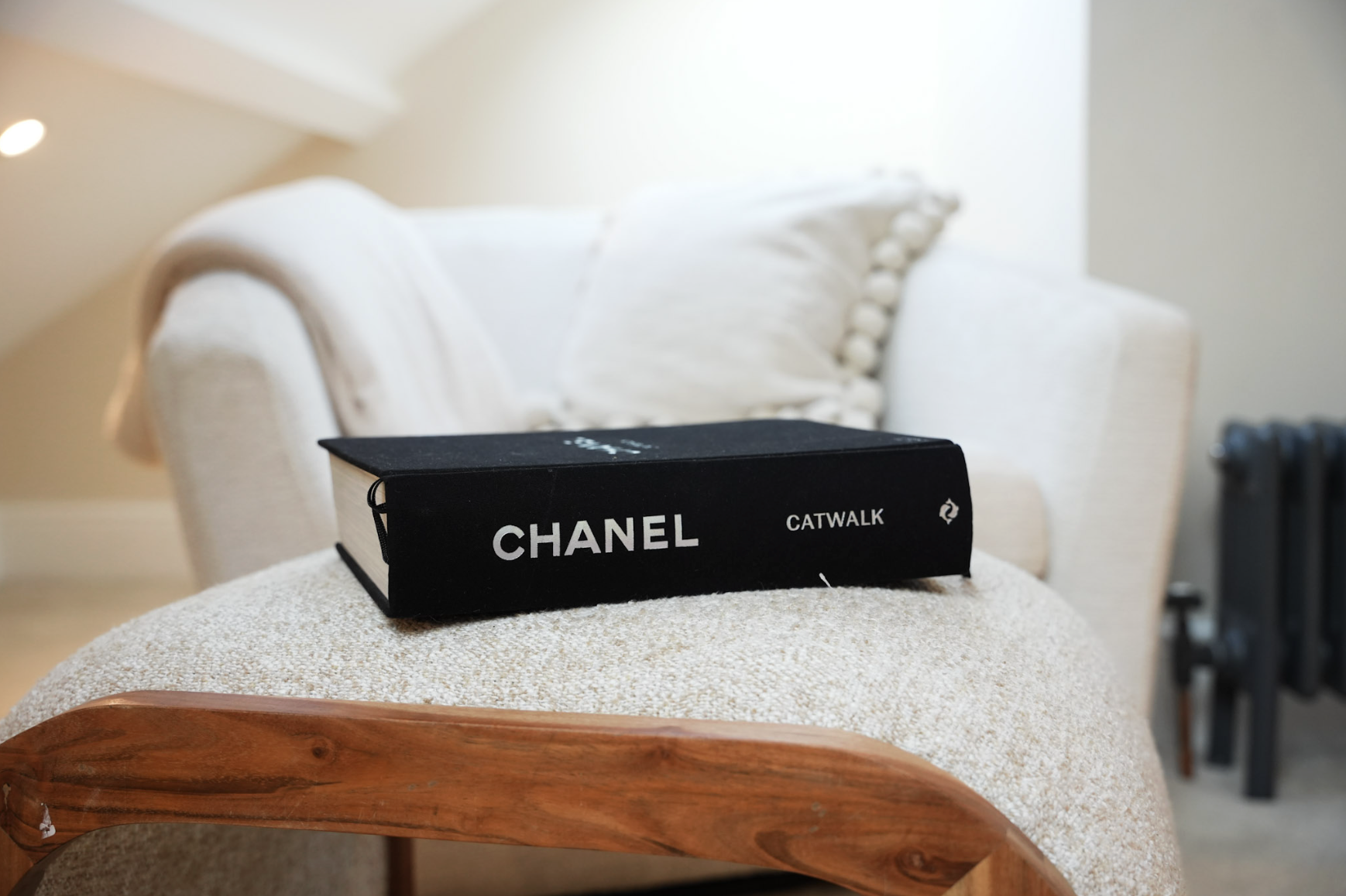 Black Chanel product box labeled "Carwalk" resting on a beige textured ottoman in a living room with a cream-colored sofa and plush pillow in the background.