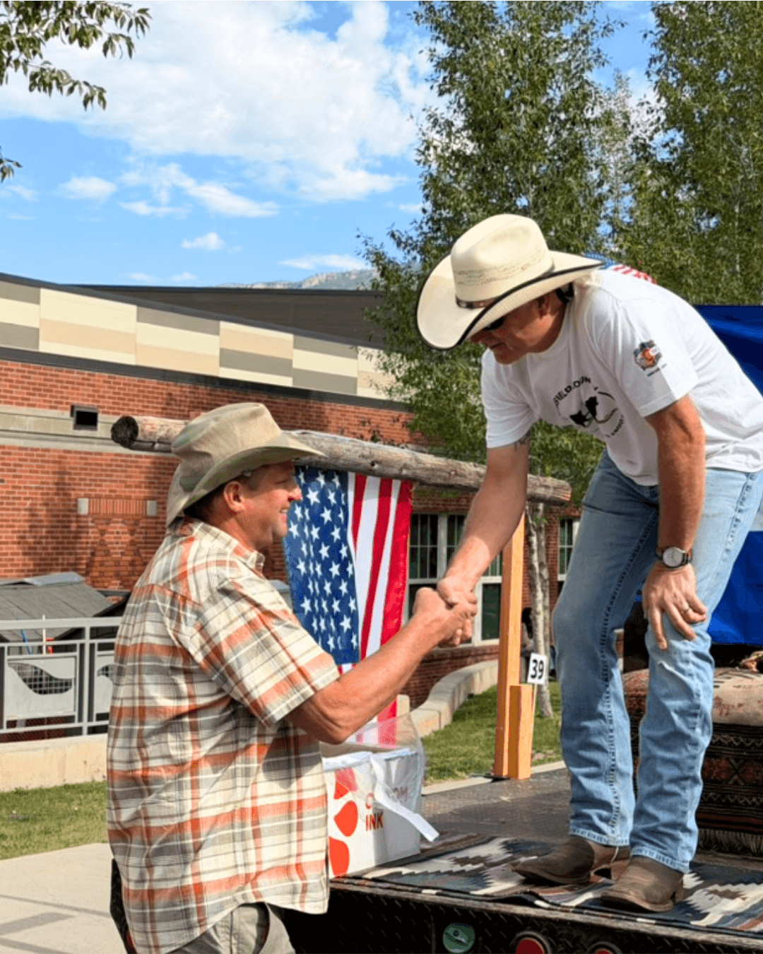 A man wearing a plaid shirt and beige cowboy hat shaking hands with a woman in a white t-shirt and large cowboy hat on a platform outdoors with a building and American flag in the background.