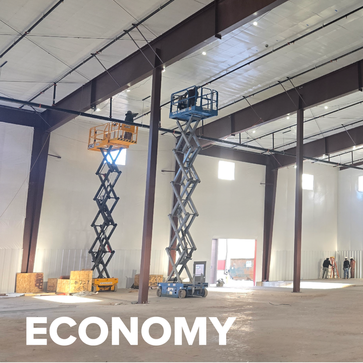 Interior of a large industrial building under construction, with two scissor lifts being used by workers, and the word 'ECONOMY' written at the bottom.