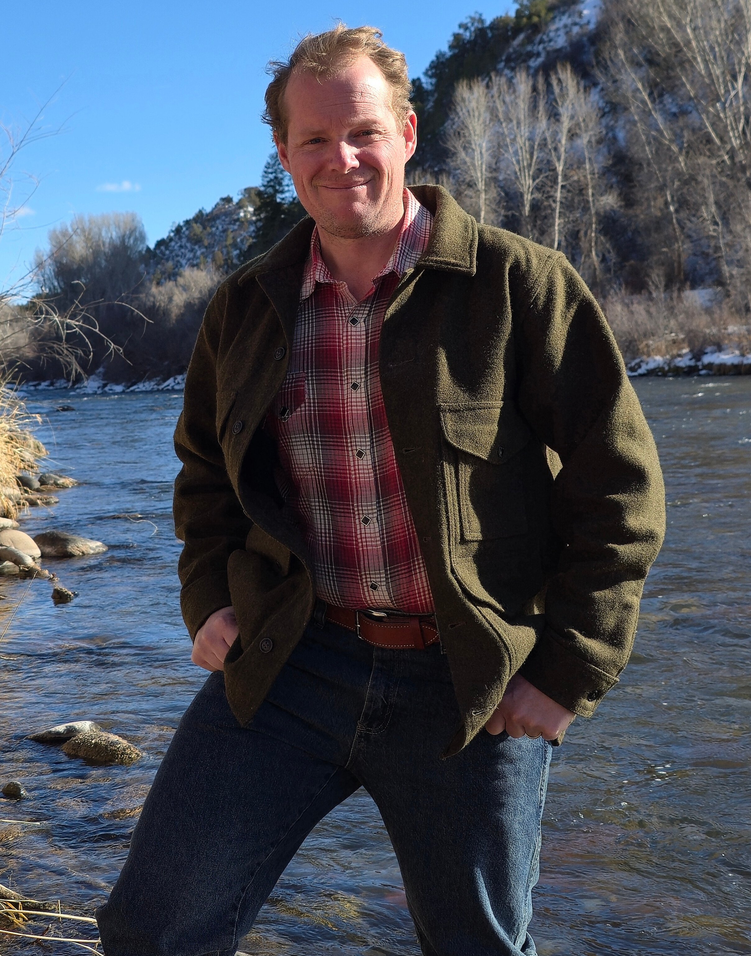 Man standing in shallow river with trees and hills in background, smiling at the camera in bright sunlight