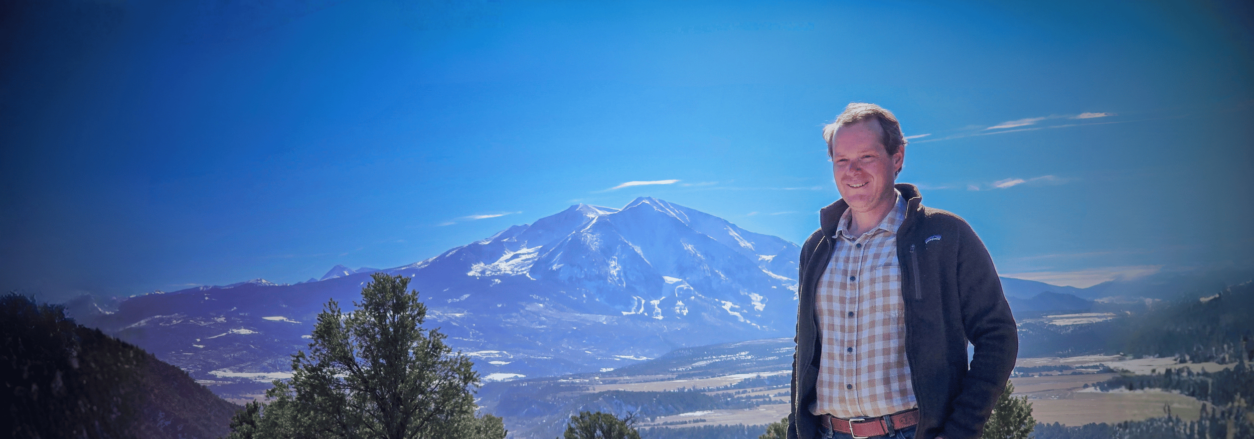 A man standing outdoors with a snow-capped mountain in the background under a blue sky.