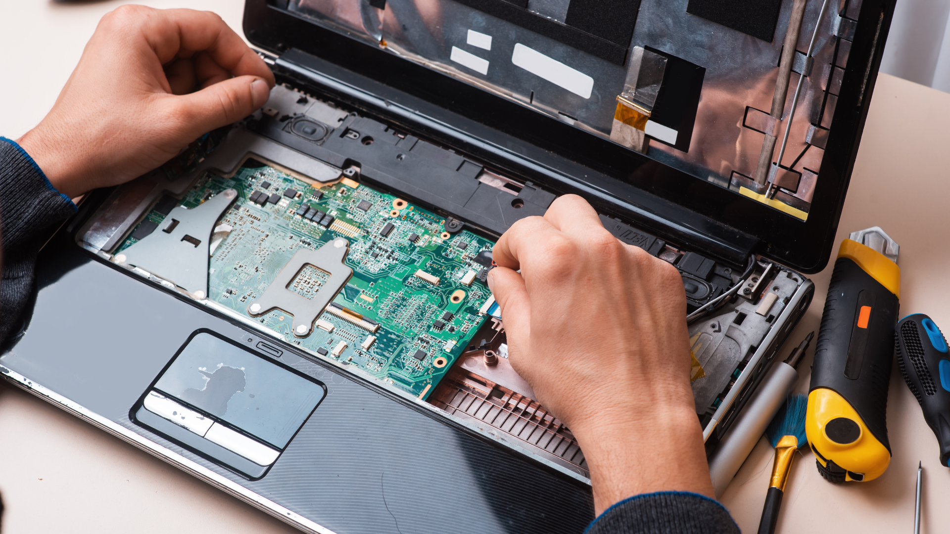 Close-up of technician repairing a laptop, with hands working inside the open device, surrounded by repair tools on a white surface.