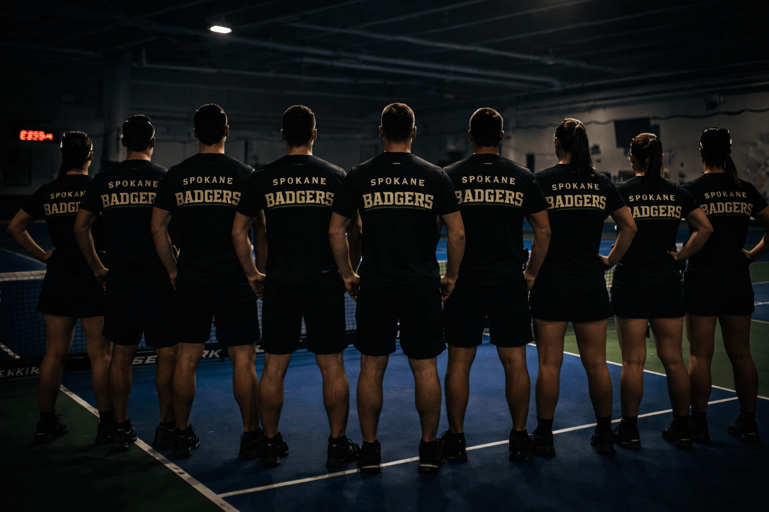A group of nine athletes, wearing black Spokane Badgers shirts, stands in a row on an indoor tennis court, facing away from the camera.
