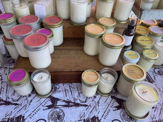 A display of various scented candles in glass jars with metal lids on a wooden table and decorative fabric underneath.
