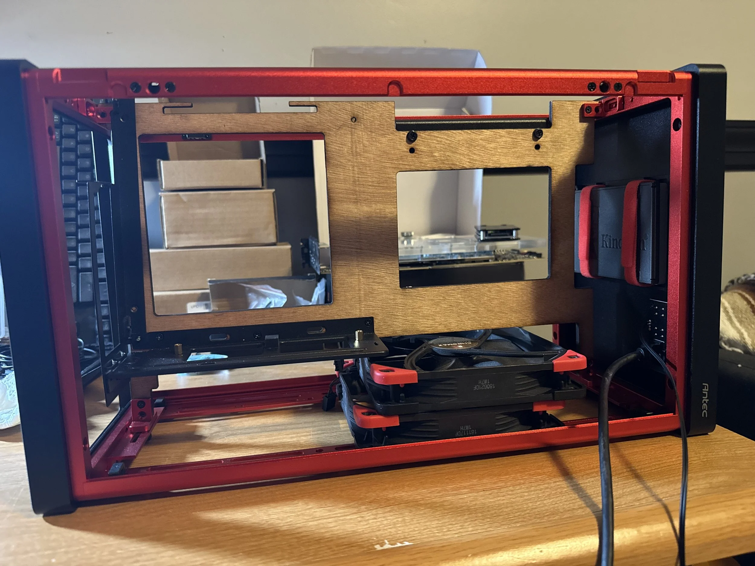 The image shows the interior of a computer case with red and black metal framing, a wooden panel in the middle, two computer cooling fans at the bottom, and some cables connected to components.