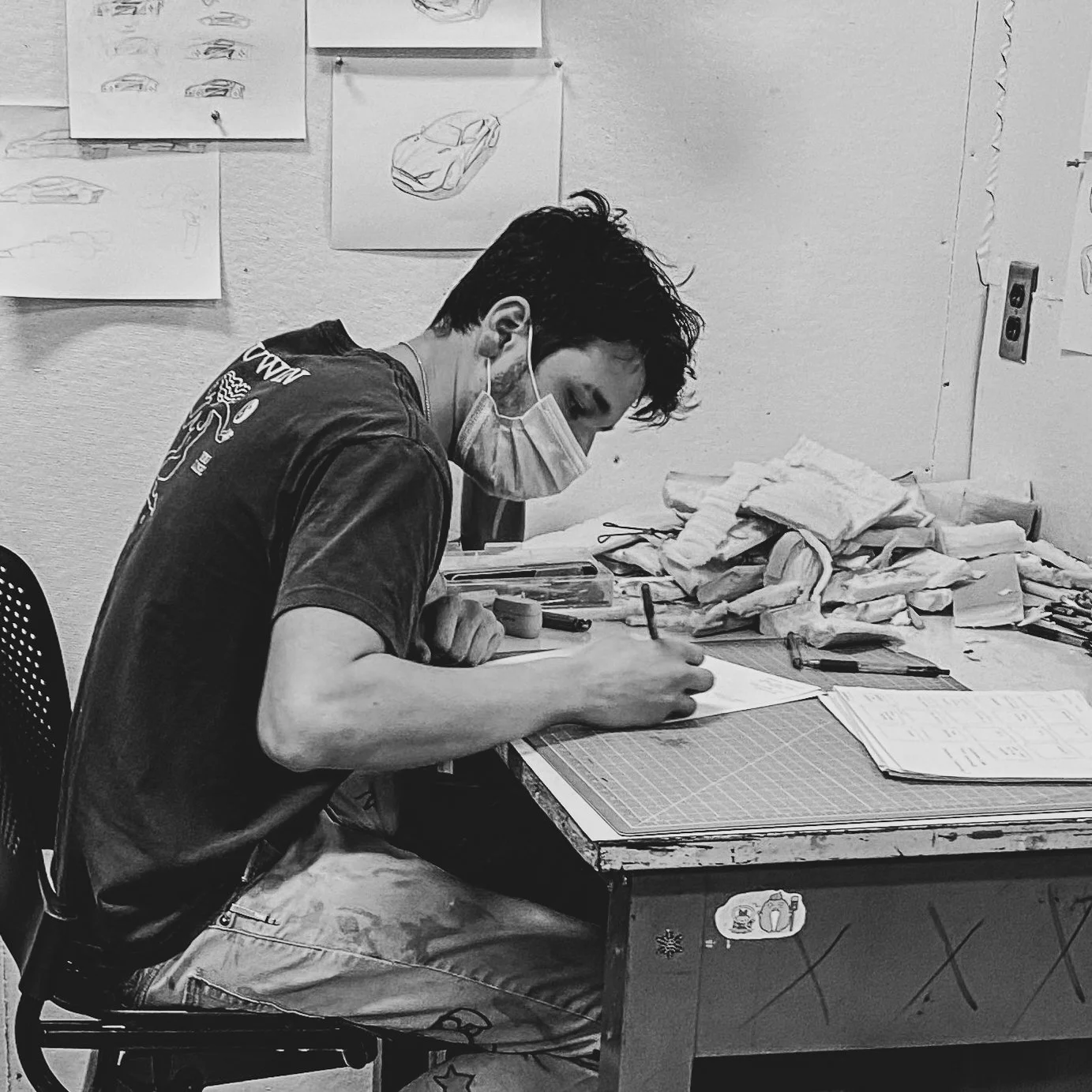 A person wearing a face mask, sitting at a cluttered desk, drawing or writing on paper. The wall behind features sketches of cars and some isometric drawings, with art supplies and papers scattered on the desk.