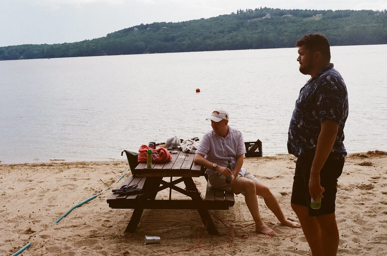 Two men on a beach near a lake, with one sitting at a picnic table and the other standing holding a drink, during daytime with silhouette of hills in the background.