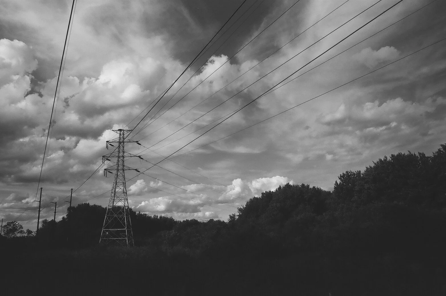 Black and white photo of power lines and transmission towers with a cloudy sky and trees in the background.