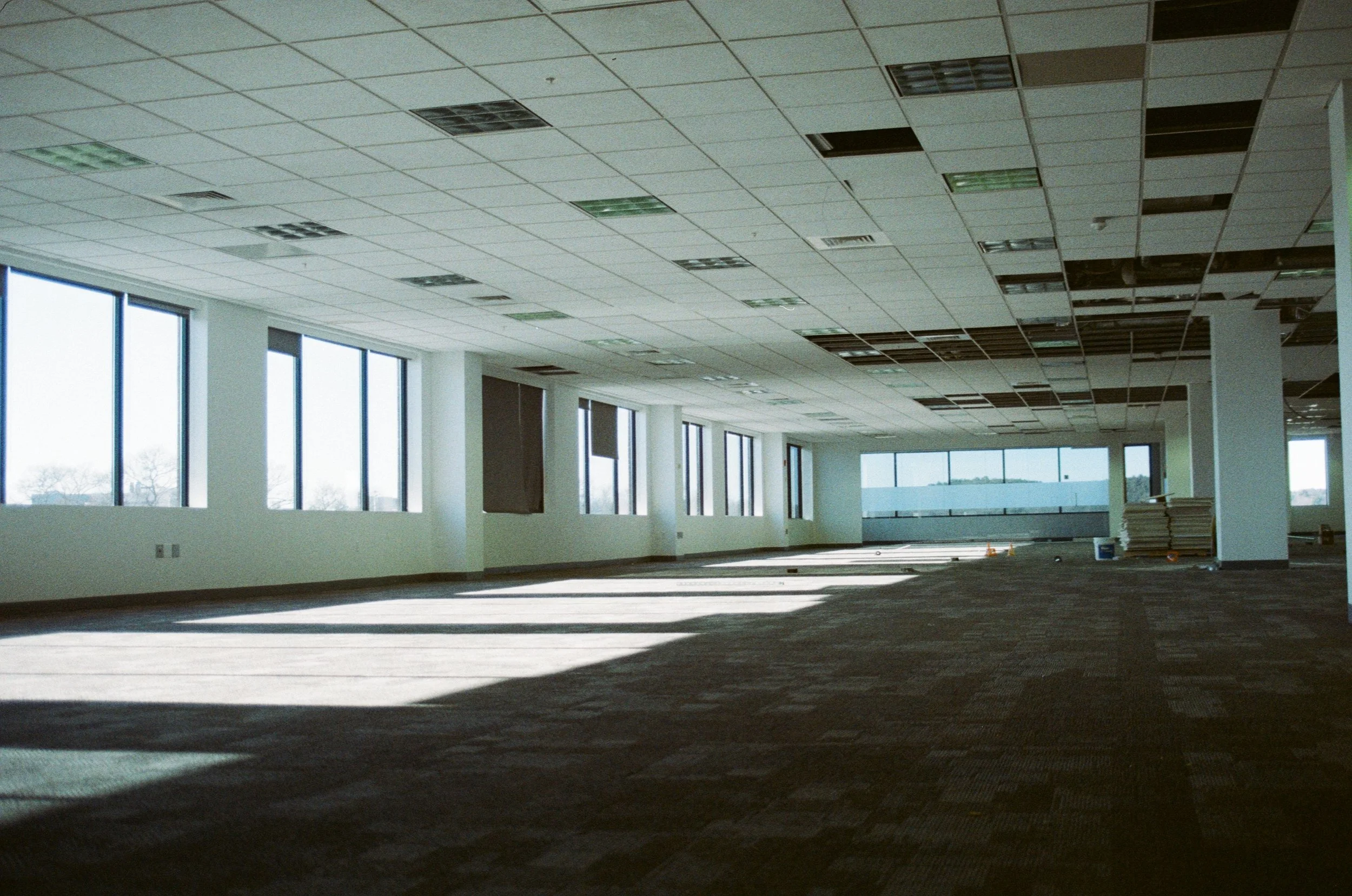 Empty office space under construction with large windows, some ceiling tiles missing, and building materials on the floor.
