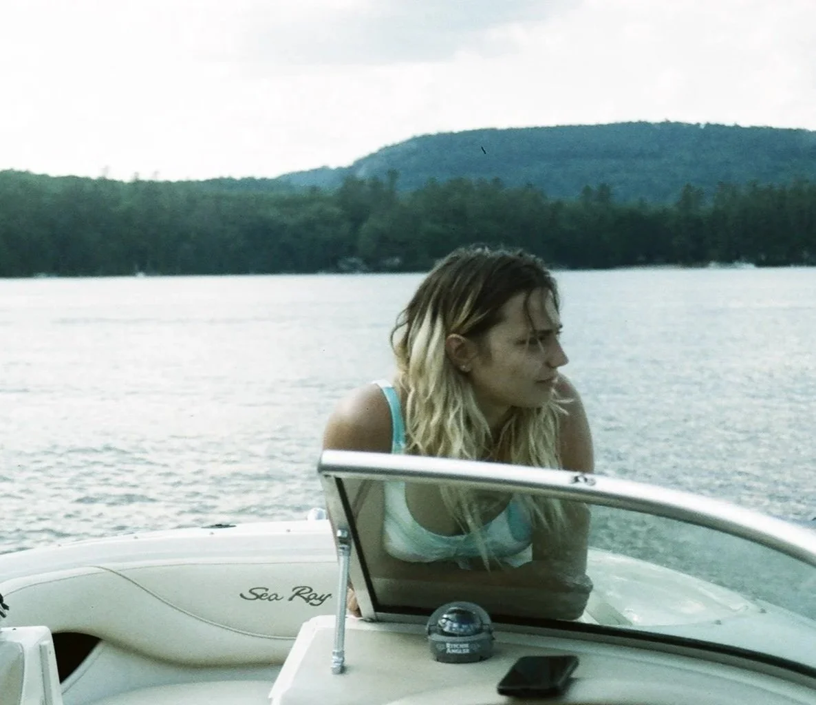A woman with blonde hair sitting on a boat named Sea Ray, looking toward the water with her arms crossed. The boat is on a lake with tree-covered hills in the background.