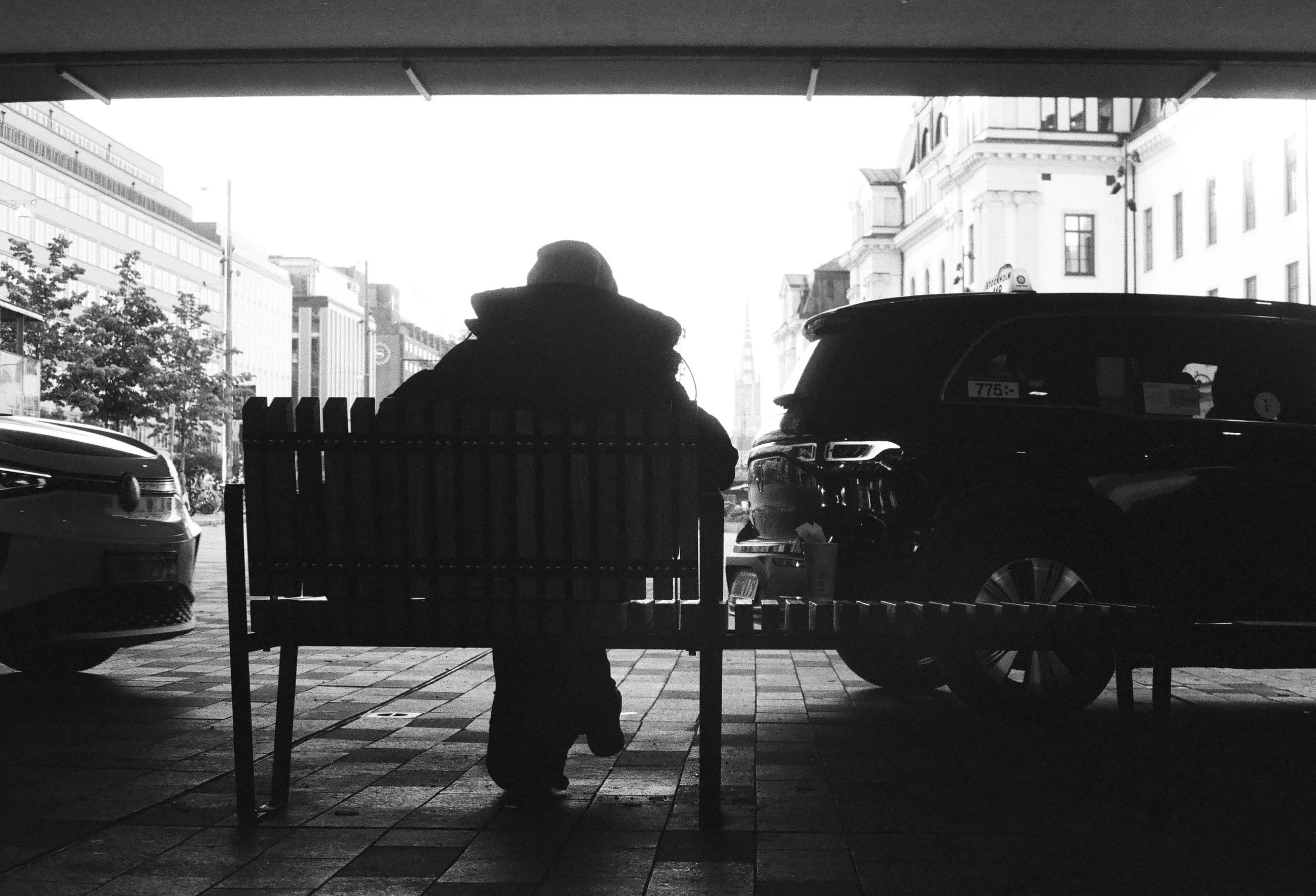 A person sitting on a park bench under a shelter facing a street with parked cars and buildings in the background.