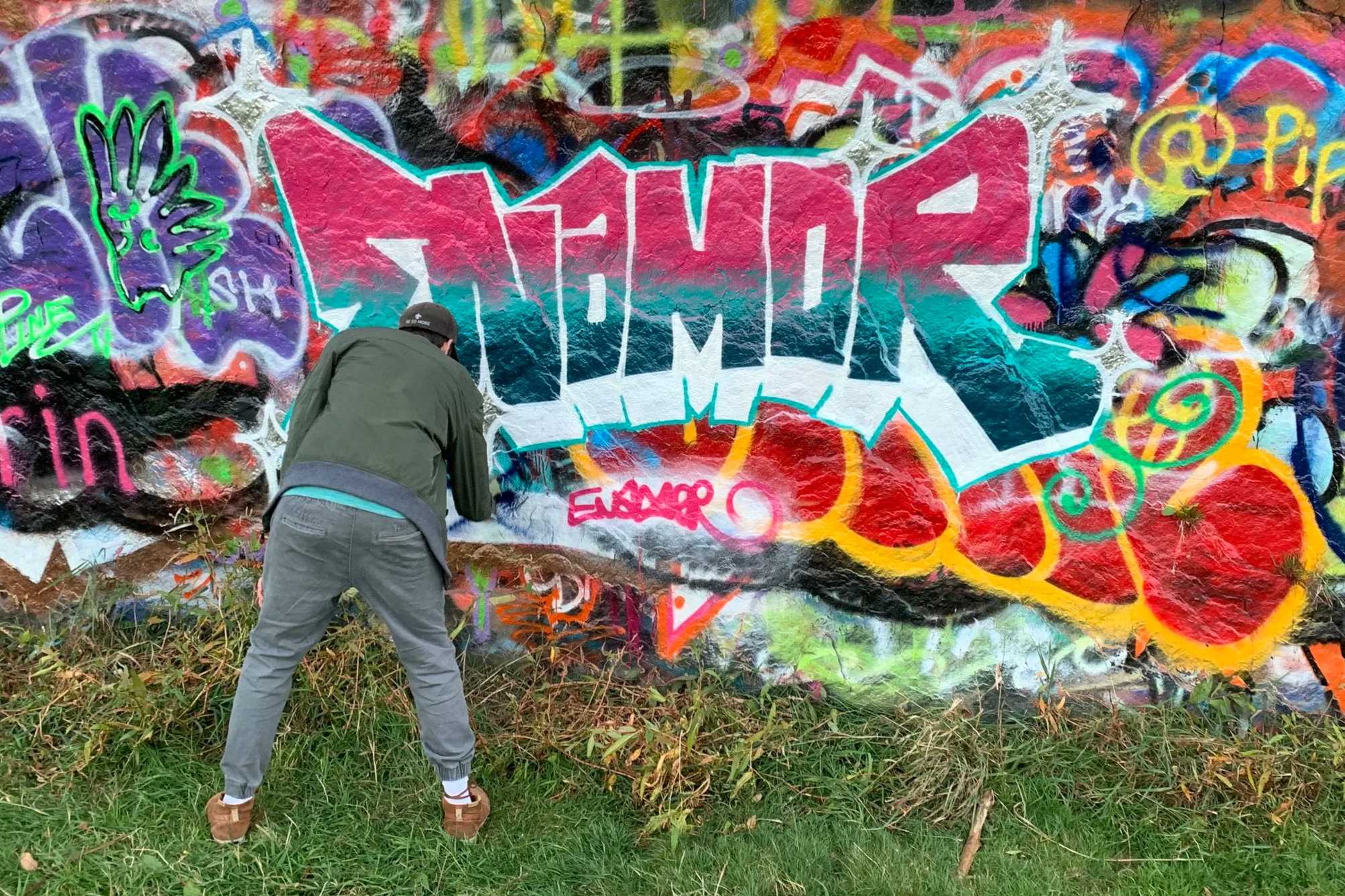 Person spray painting graffiti on a colorful wall with various tags, images, and vibrant designs.