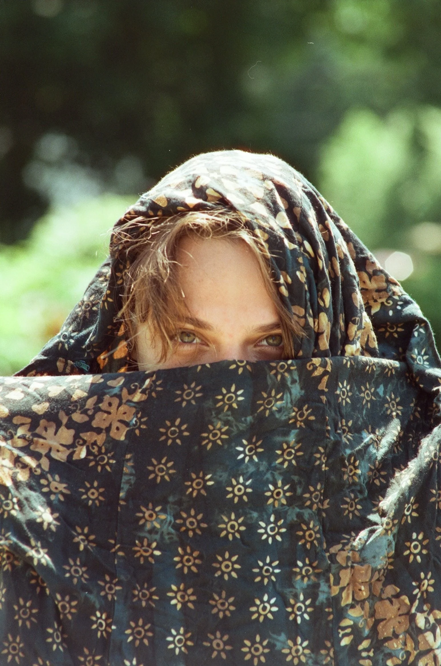 Person with striking green eyes and light brown hair, partially covered with a dark patterned fabric, outdoors with blurred green foliage in the background.