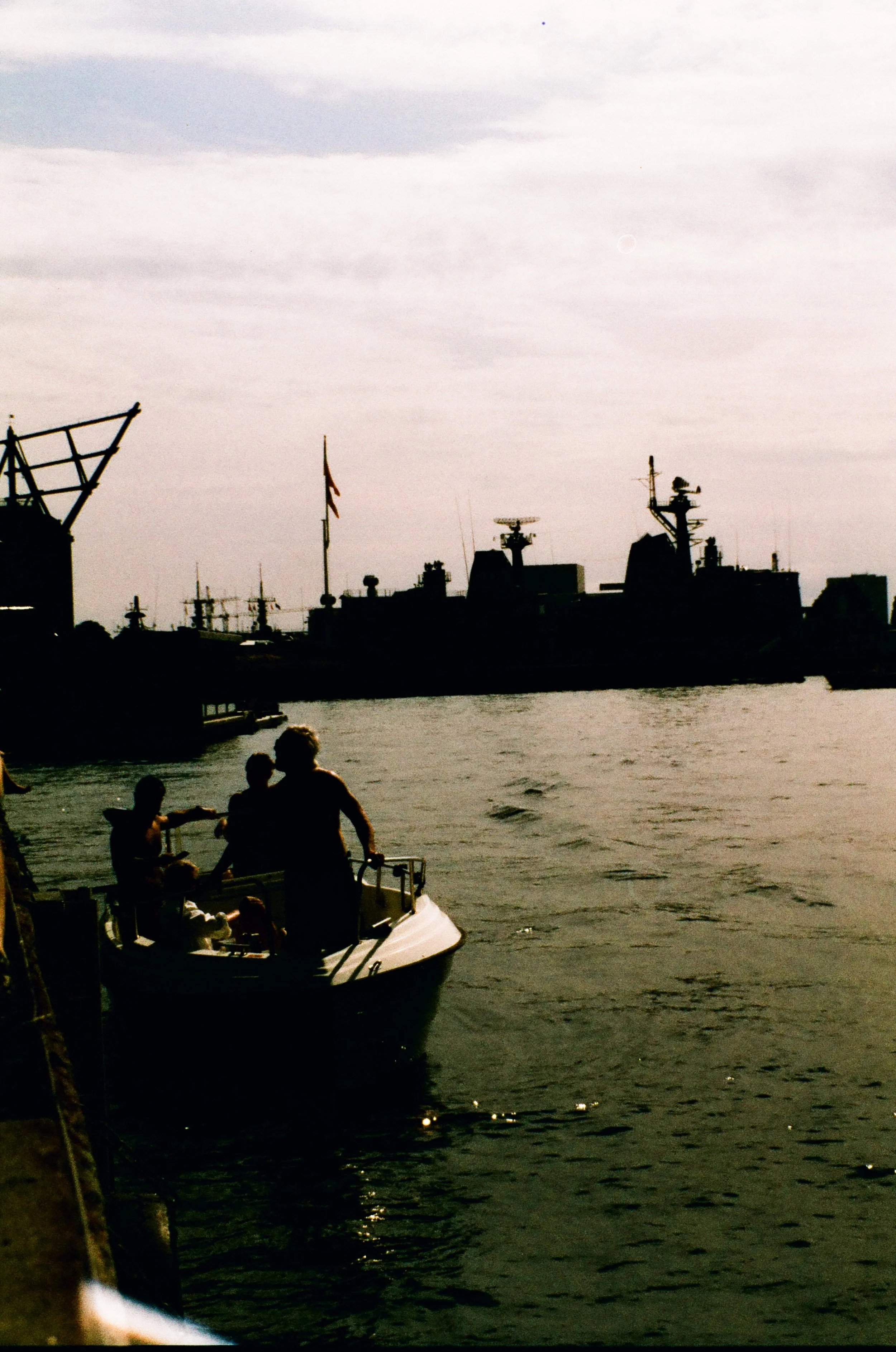 People on a boat near a dock with ships and naval vessels in the background.