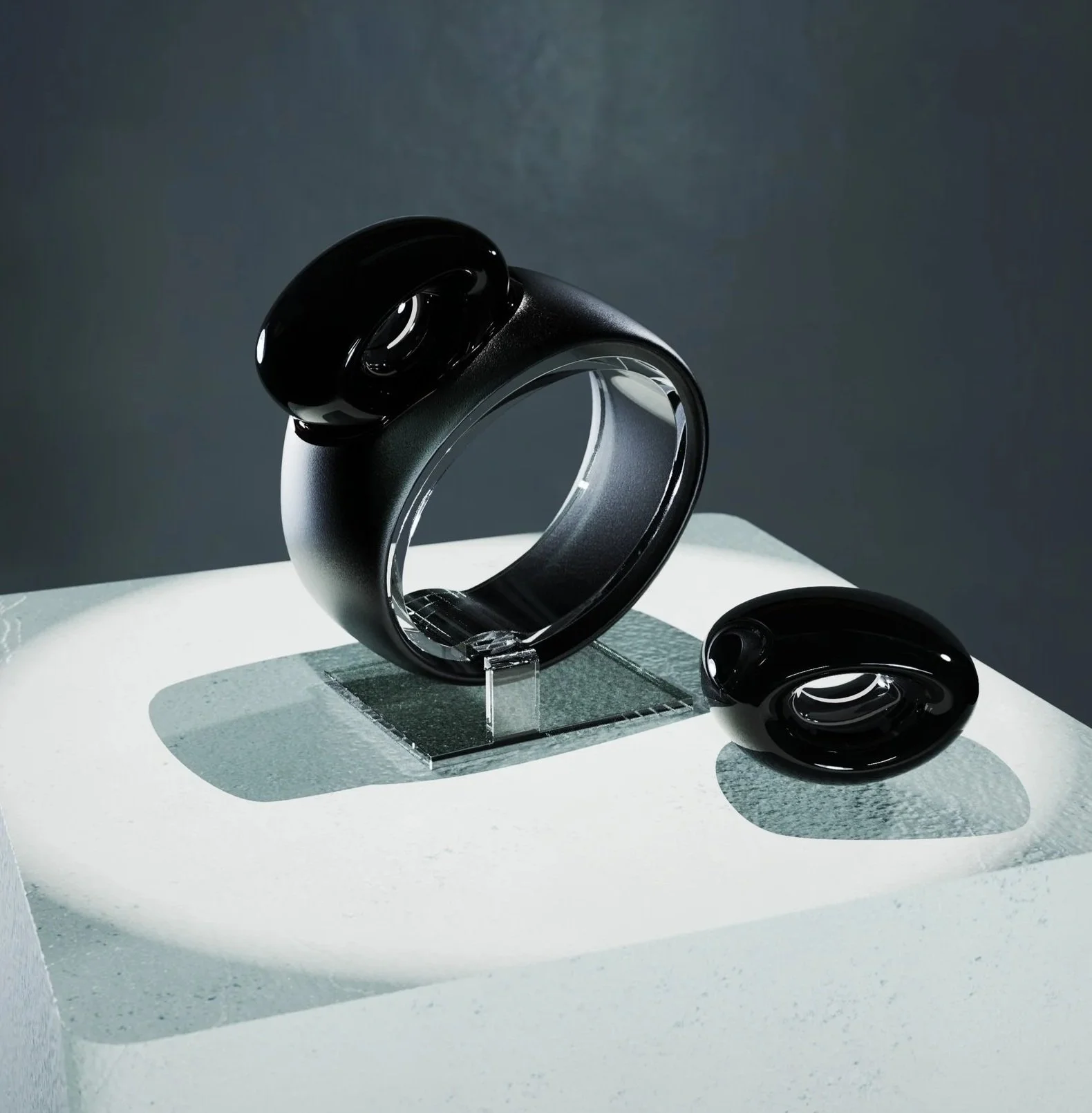 A black ring with a large, oval, glossy black stone on top, displayed on a clear stand, with a matching black stone accessory nearby, against a gray background.