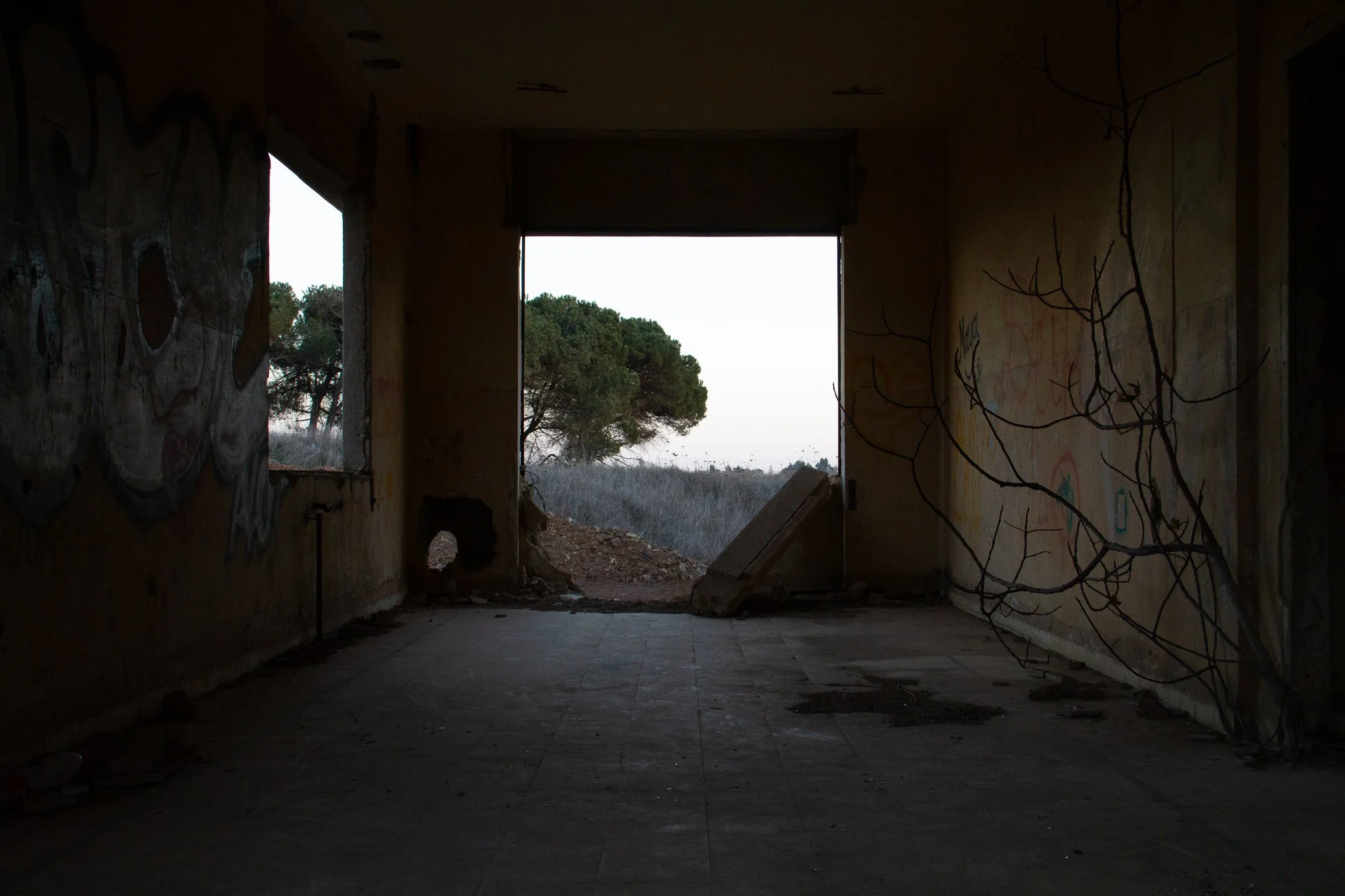 An abandoned room with graffiti walls, broken window, a collapsed wall section, and a view of trees and open sky outside.