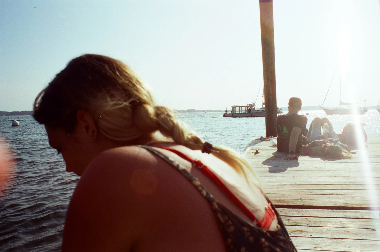 A woman with blonde hair in a braid sitting near the water on a dock, with boats in the background and sunlight reflecting on the water.