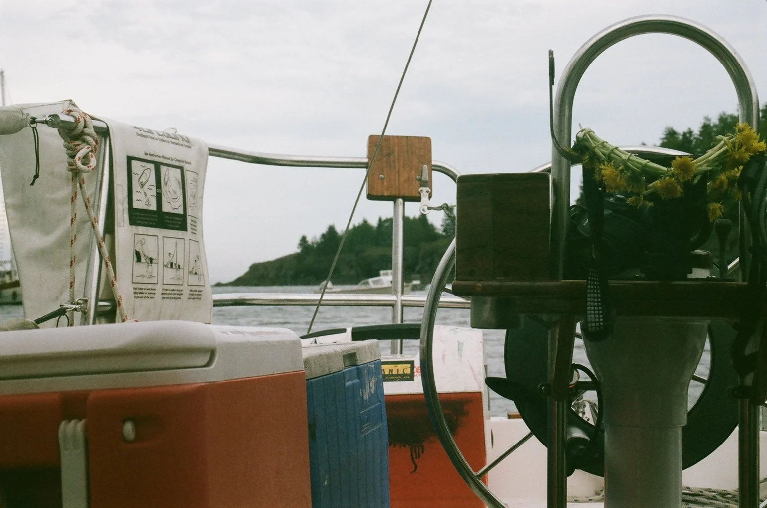 Close-up of a sailboat's deck featuring a steering wheel with a yellow floral wreath hanging on it, a wooden dock, and water with a distant landmass in the background under a cloudy sky.