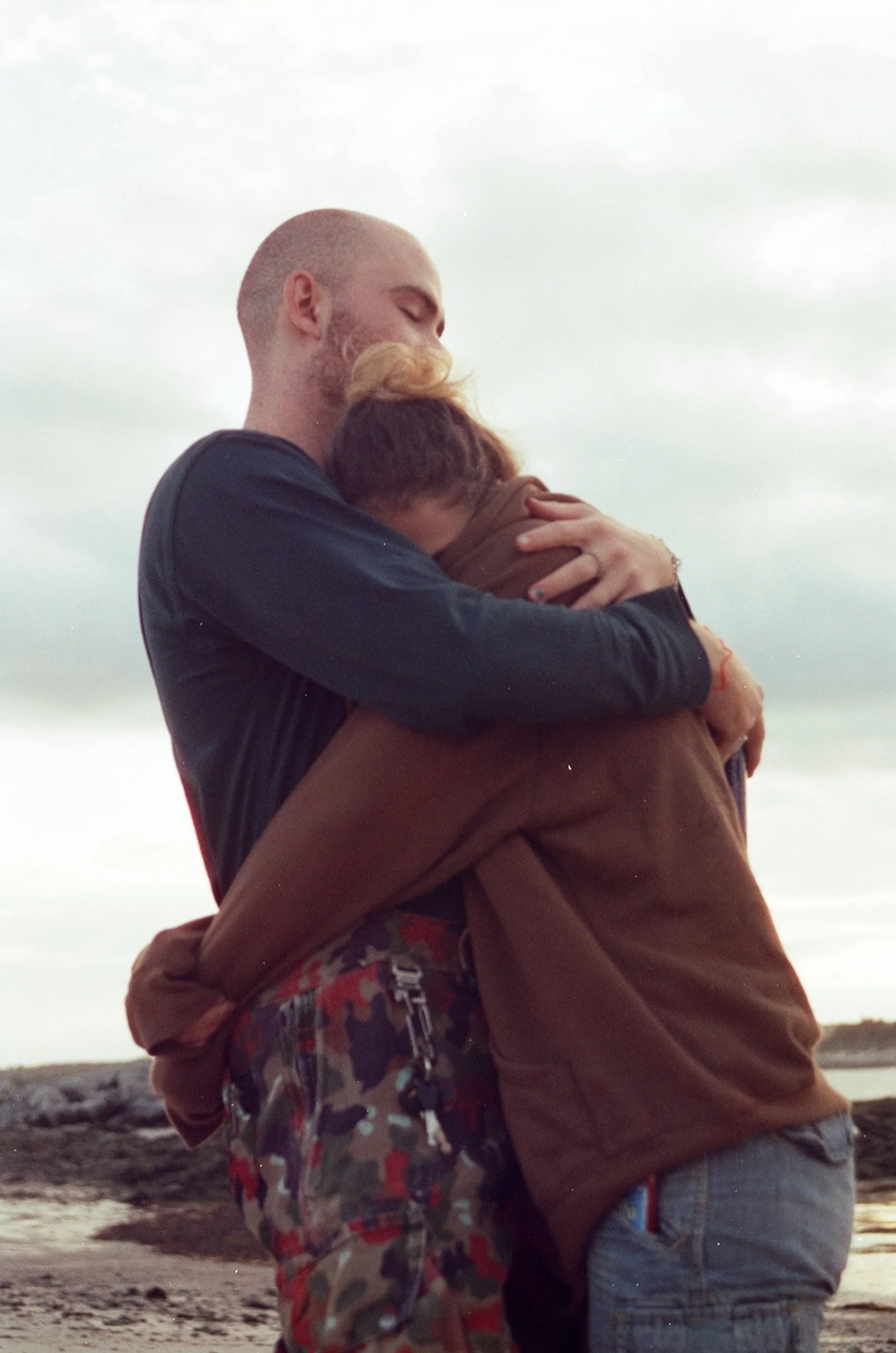 A couple embracing outdoors, with the woman hugging the man and both appearing emotional, against a cloudy sky and rocky landscape.
