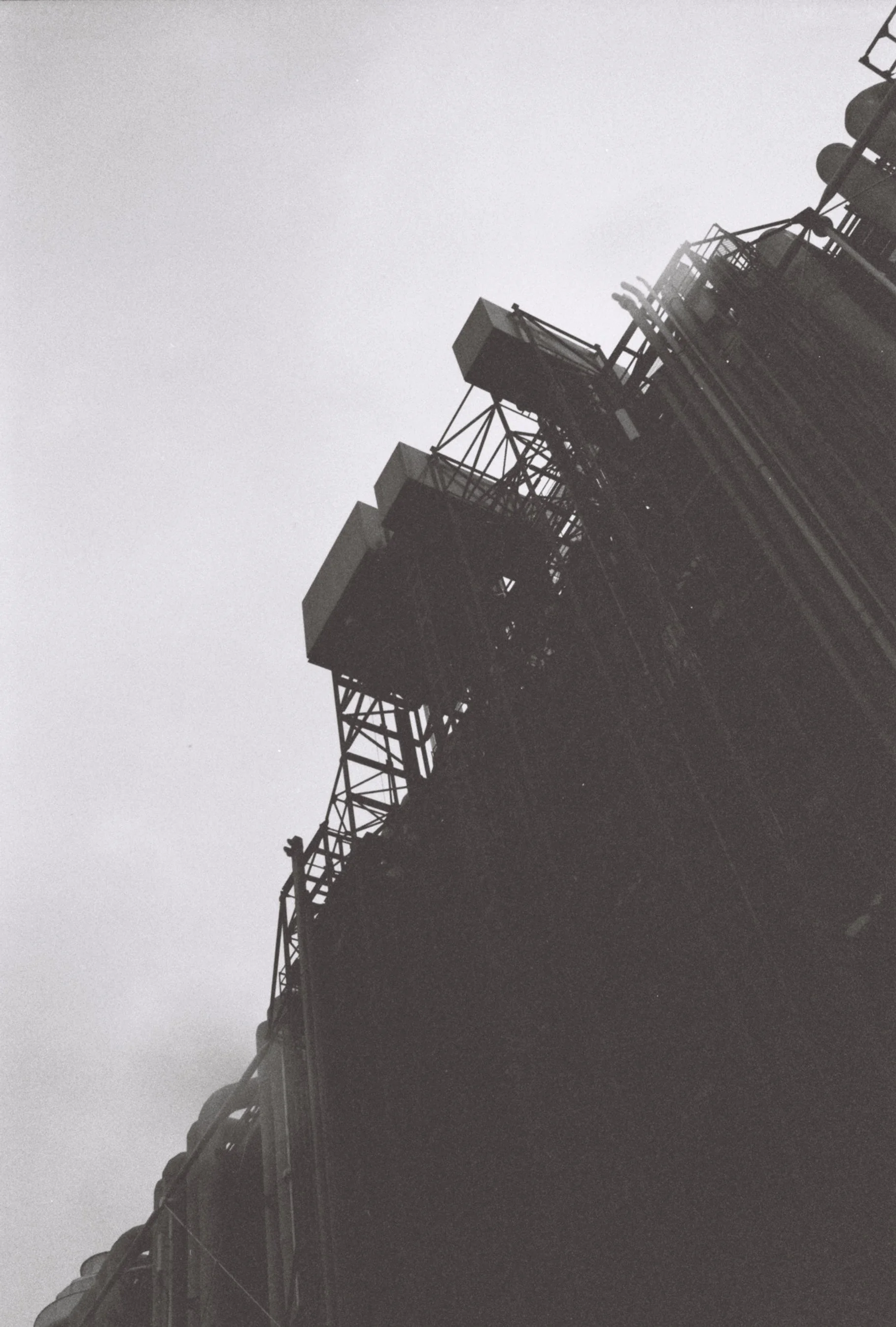 Black and white photo of a building's rooftop with industrial structures and machinery, shot from a low angle against a cloudy sky.