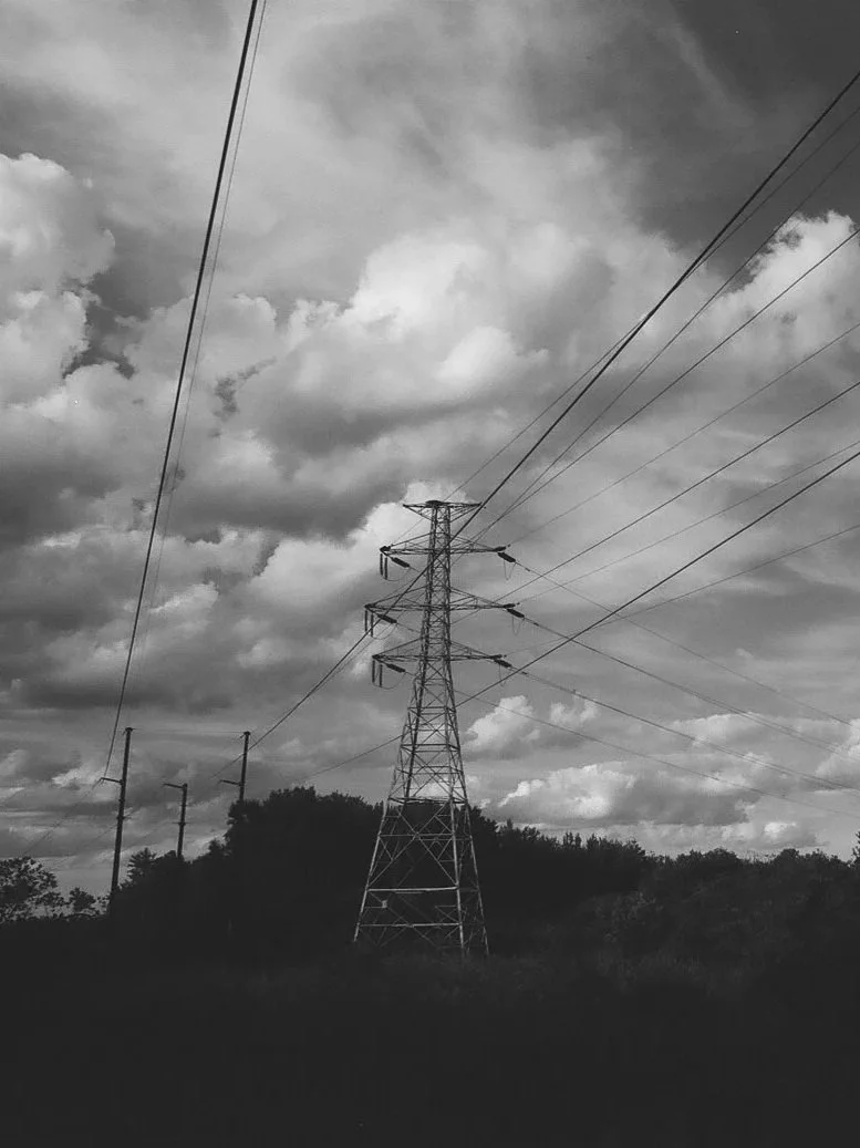 Black and white photo of power lines and a transmission tower against cloudy sky and trees in the background.