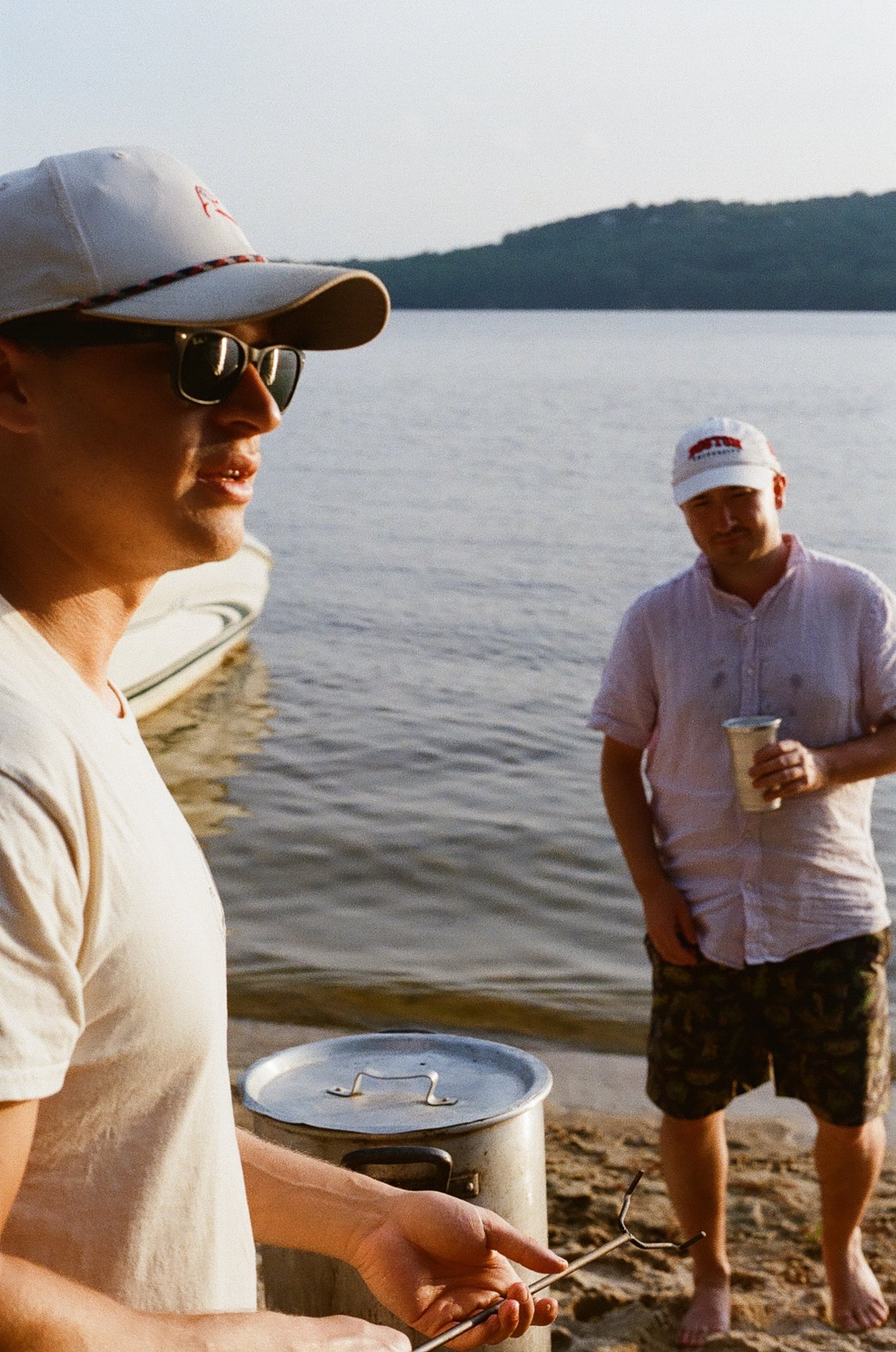 Two men stand near a lake or river at sunset, with one holding a butter knife and the other holding a drink, a boat nearby, and a wooded hill in the background.