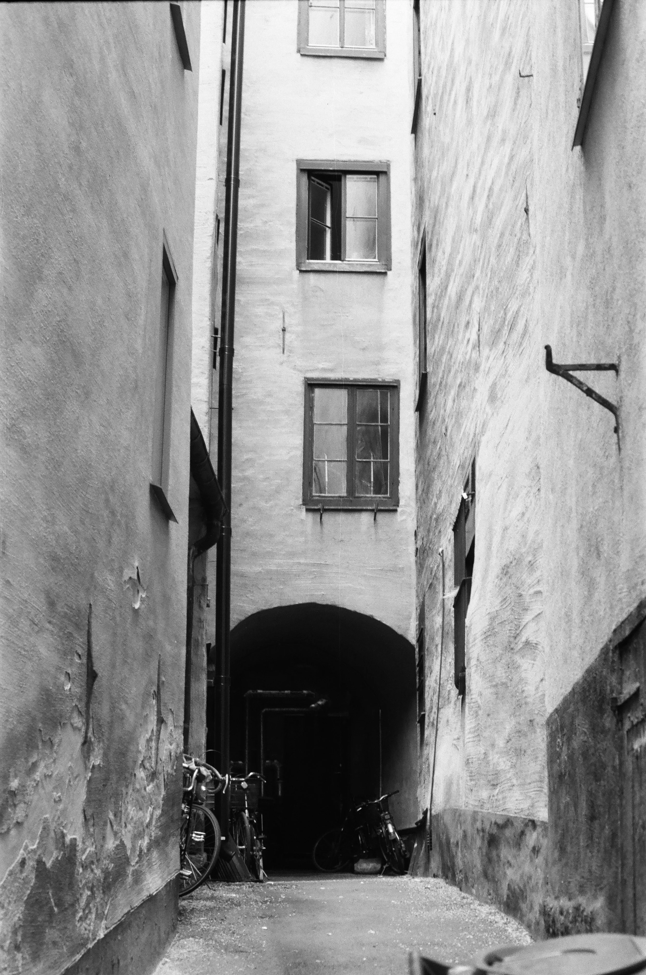 Black and white photo of a narrow alleyway with old buildings, windows, bicycles, and an arched passageway at the end.