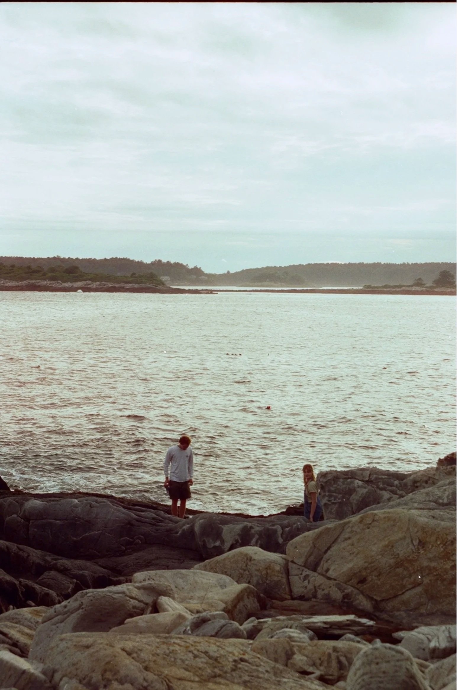 Two people standing on rocks by the water at a coastal landscape, overcast sky