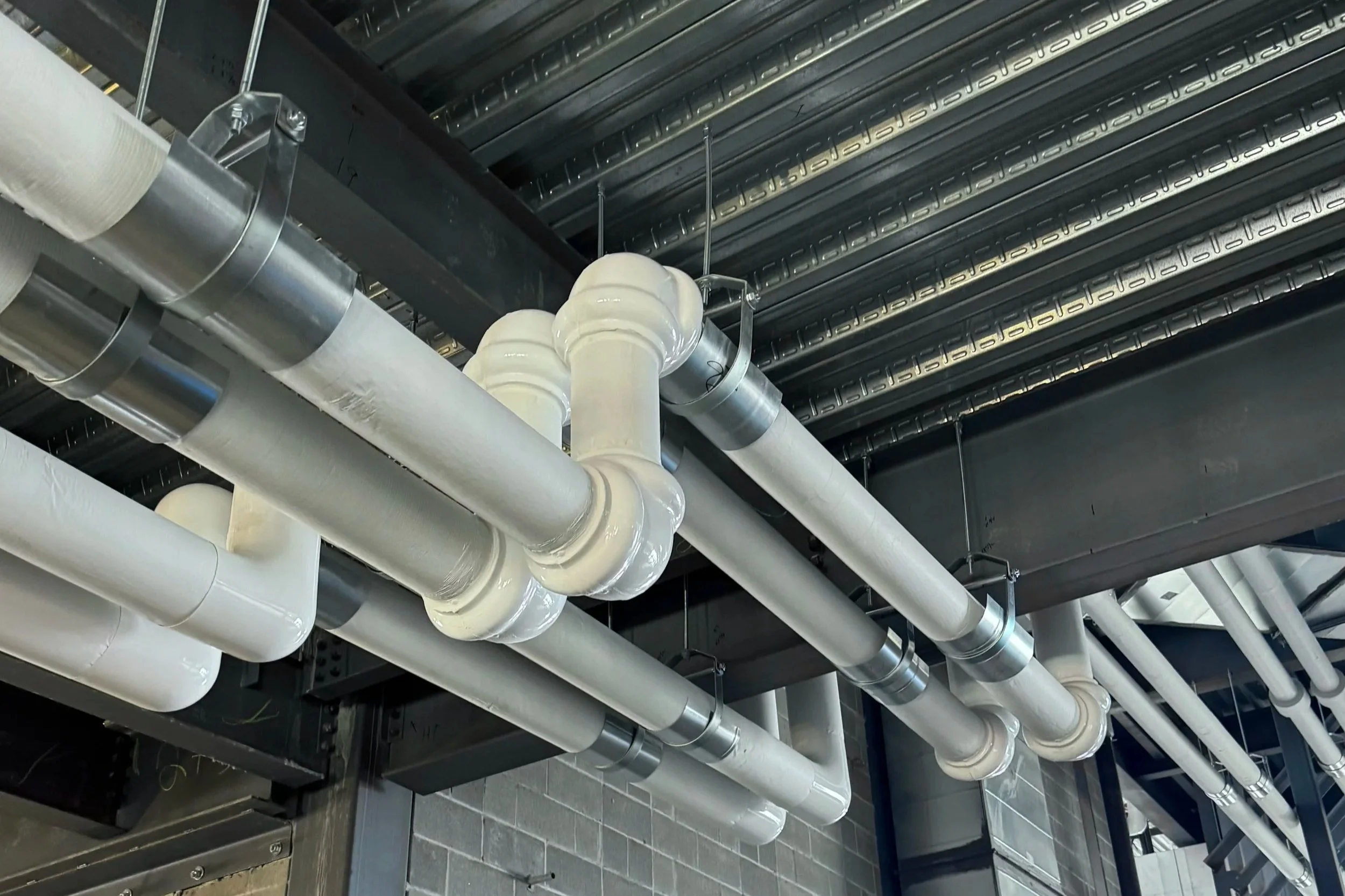Industrial ceiling with large white and metallic pipes running parallel and intersecting, suspended beneath black structural beams.