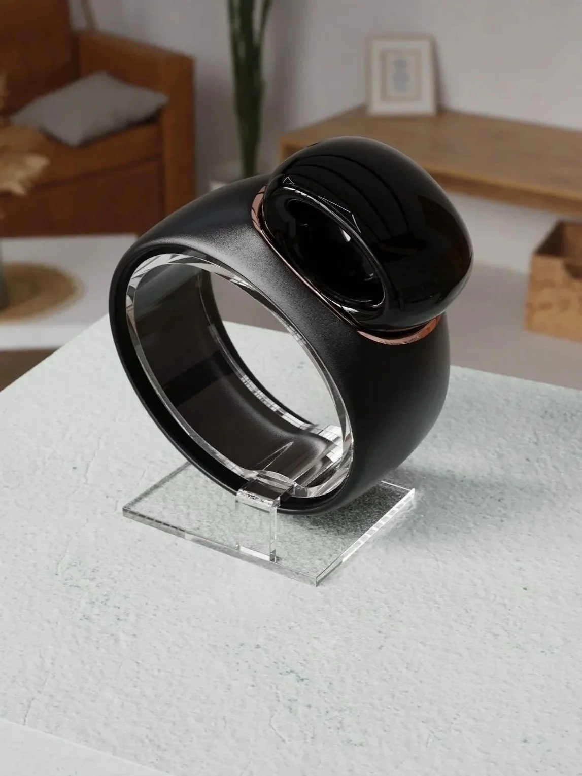 A modern black and silver wearable device, possibly a smartwatch or health tracker, displayed on a clear stand on a textured white surface in a contemporary interior setting with wooden furniture and framed art on the wall.