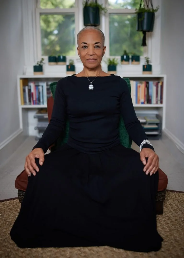 A woman with short gray hair sitting on a low ottoman in a room with white walls, a window, and a bookshelf. She is wearing a black long-sleeved top, and has a calm expression.