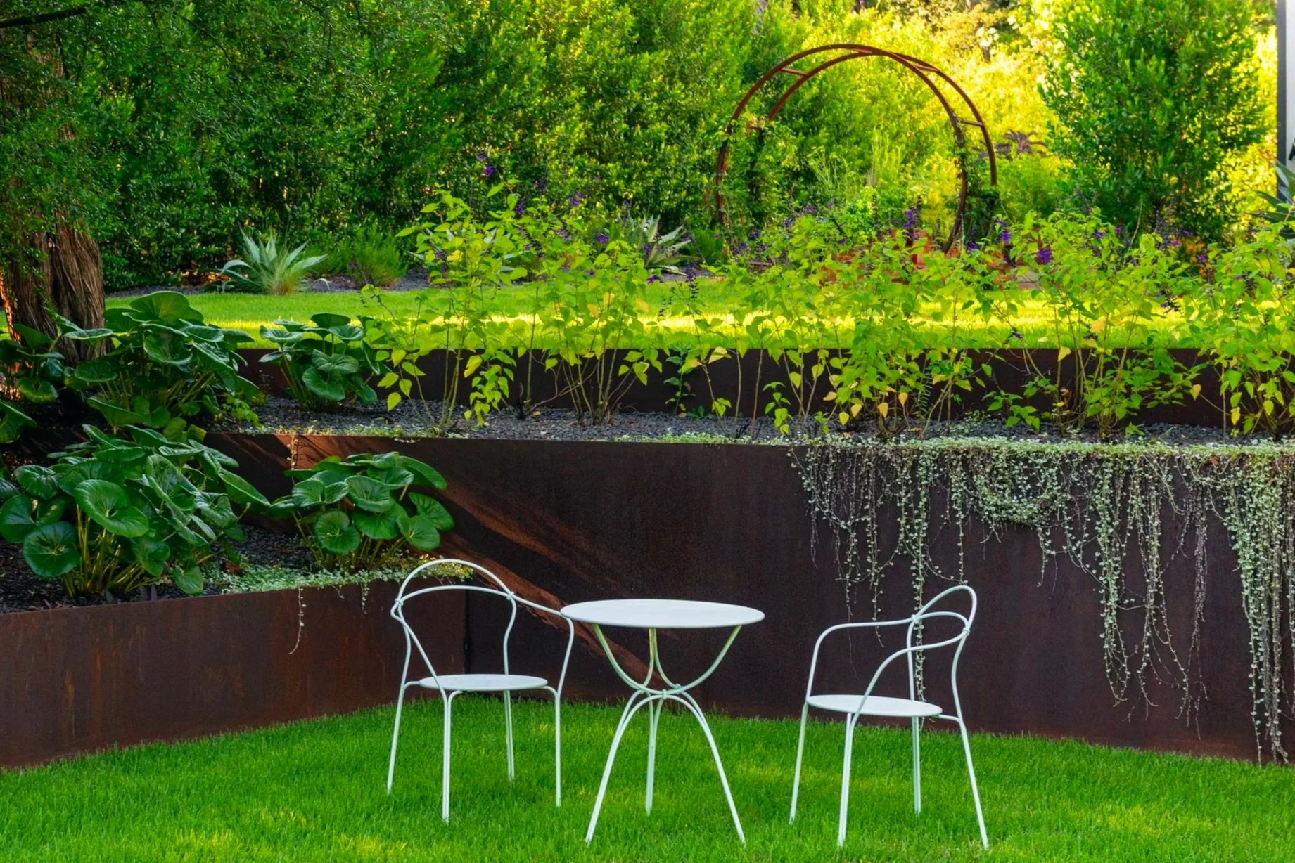 A small Fermob seating area anchors the substantial steel corner, creating an intimate, private place to gather beneath the moon gate entrance to the garden above.