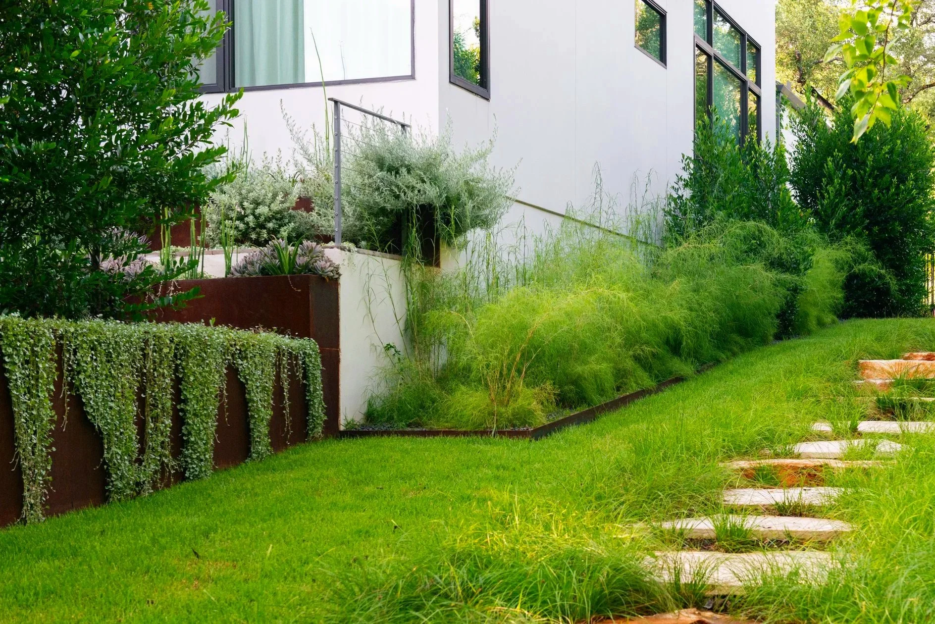 A new walkway of oversized limestone pancake boulders, softened by swaths of sedges, creates a welcoming path from the street to the home’s rear office entrance, with dramatic, layered plantings framing the journey on both sides.