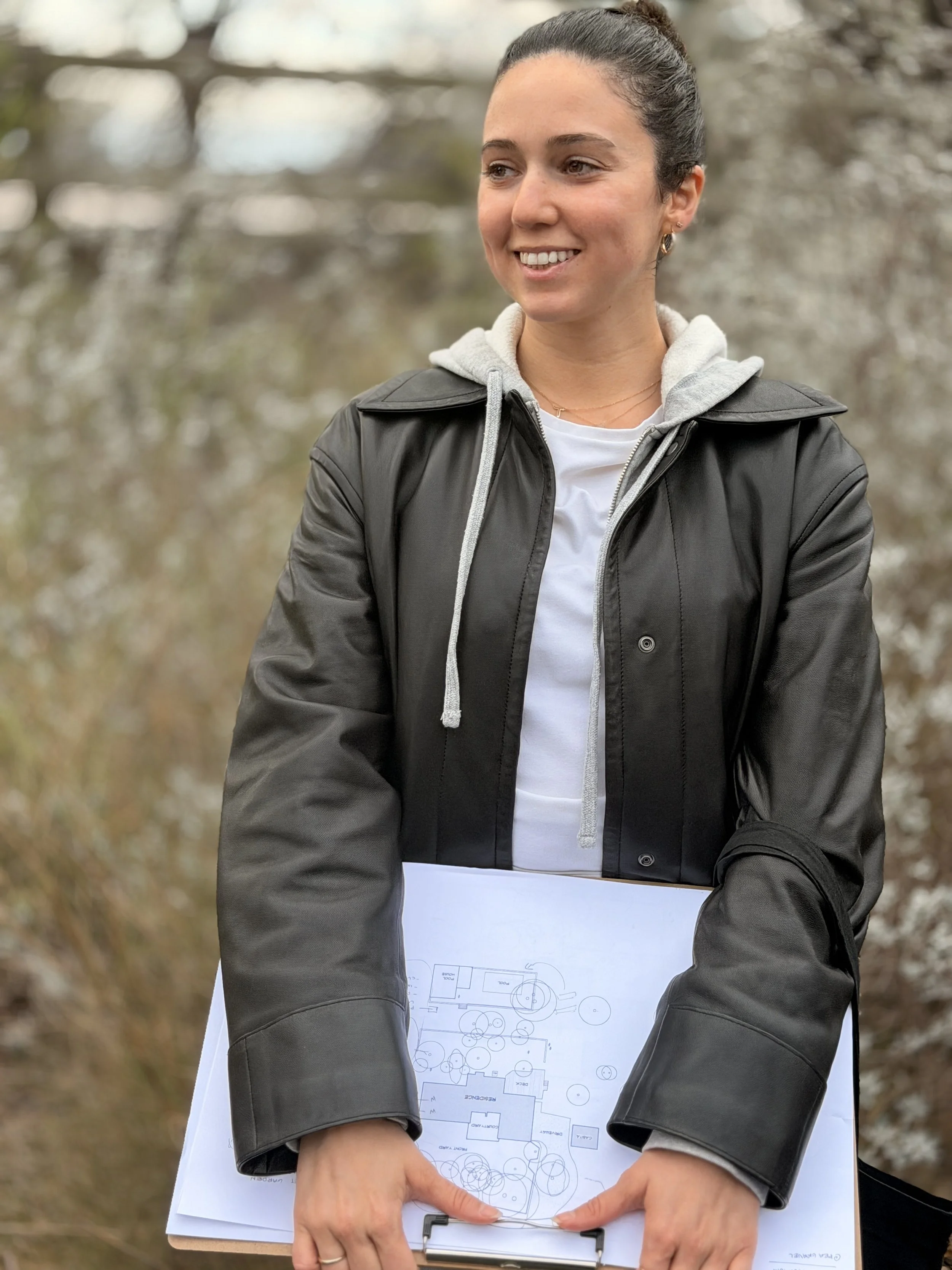 A woman with dark hair tied back, smiling, holding a clipboard with a detailed construction or landscape plan, wearing a gray hoodie under a black jacket, standing outdoors with trees in the background.