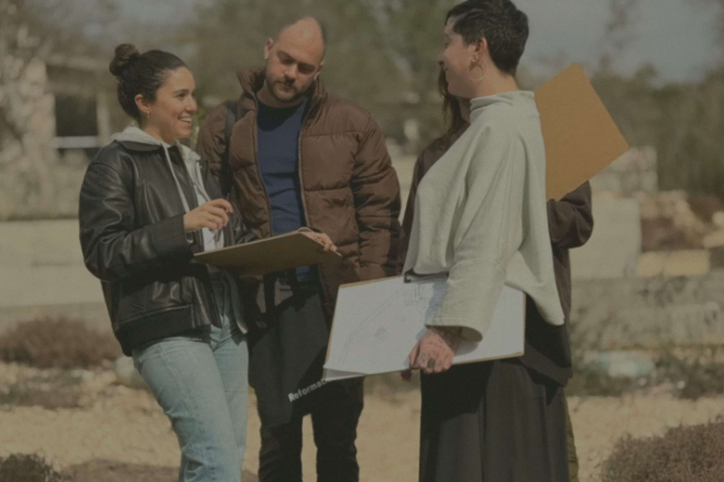 Four people standing outdoors, engaged in a conversation, with three women and one man. The woman on the left is smiling, wearing a black leather jacket and holding a clipboard. The man next to her has a beard, is bald, and is wearing a brown jacket. The woman at the right is holding a tablet and is wearing a light-colored hoodie. In the background are trees and a partly cloudy sky.