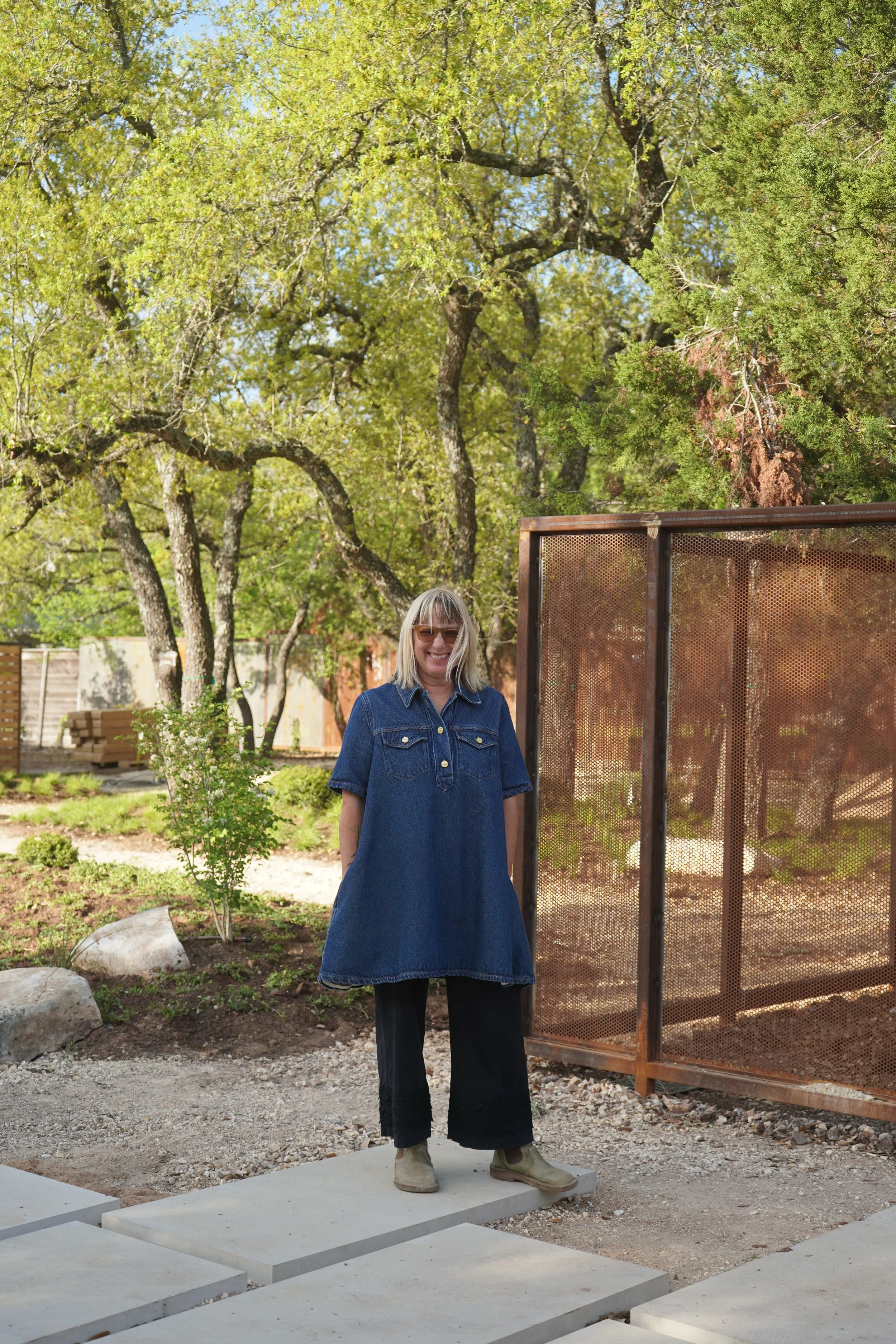 A woman stands outdoors on concrete blocks in a garden with trees, wearing a denim dress, sunglasses, and beige boots, smiling at the camera.