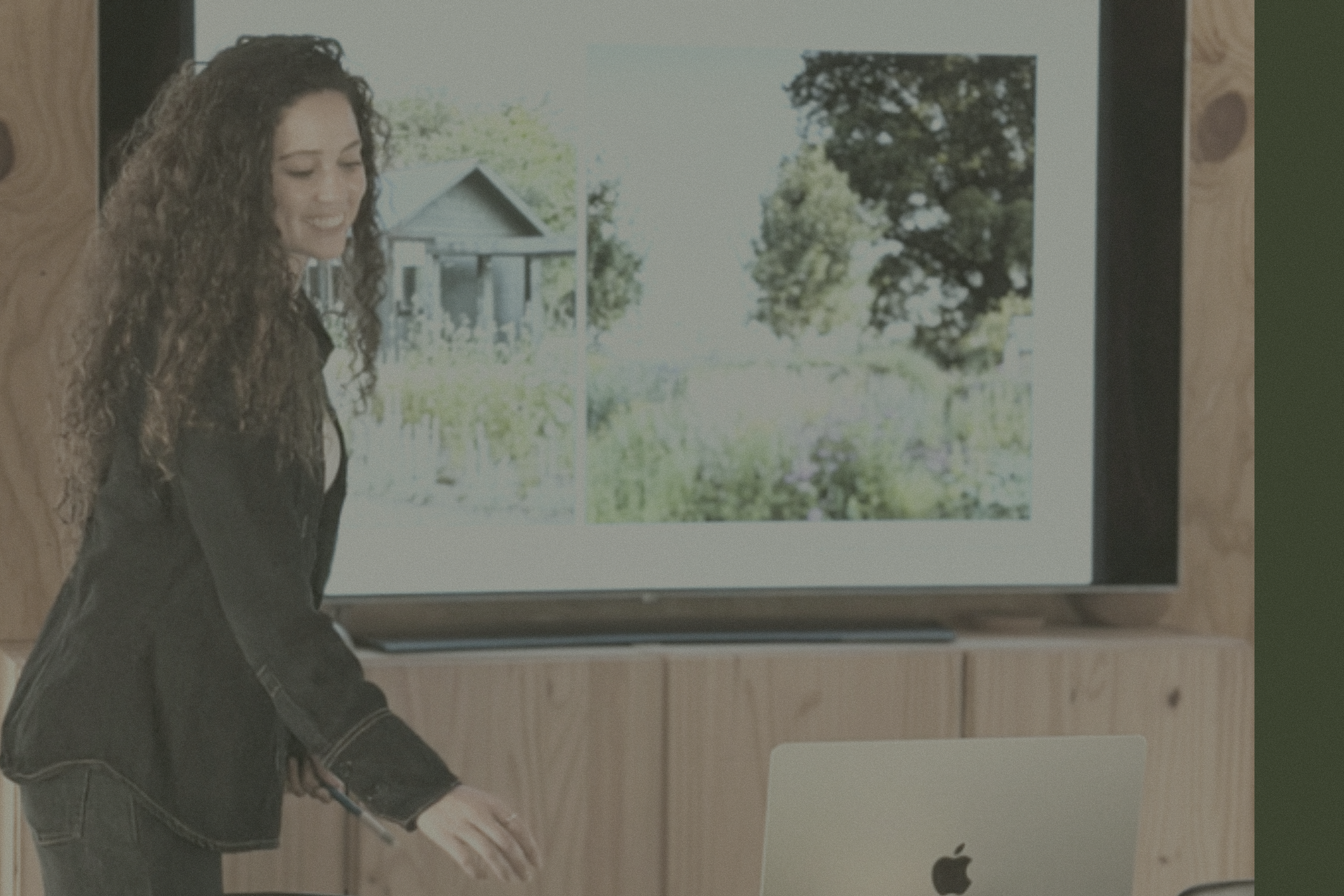 A woman with curly hair looking at a large screen displaying outdoor scenery, with an Apple computer on a wooden table in front of her.