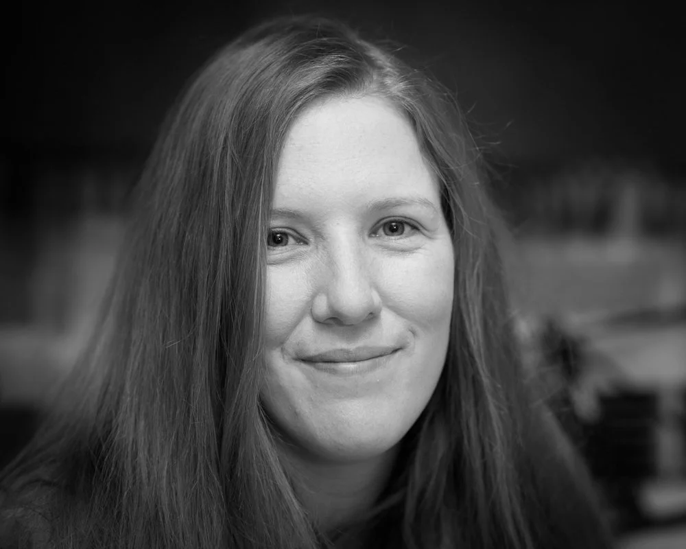Black and white portrait of a woman with long hair, smiling gently, indoors with blurred shelving in the background.
