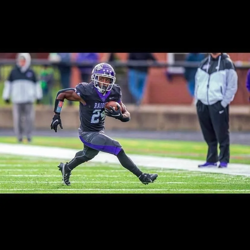 A young football player in black and purple uniform running with the ball on the field, with spectators and coaches in the background.
