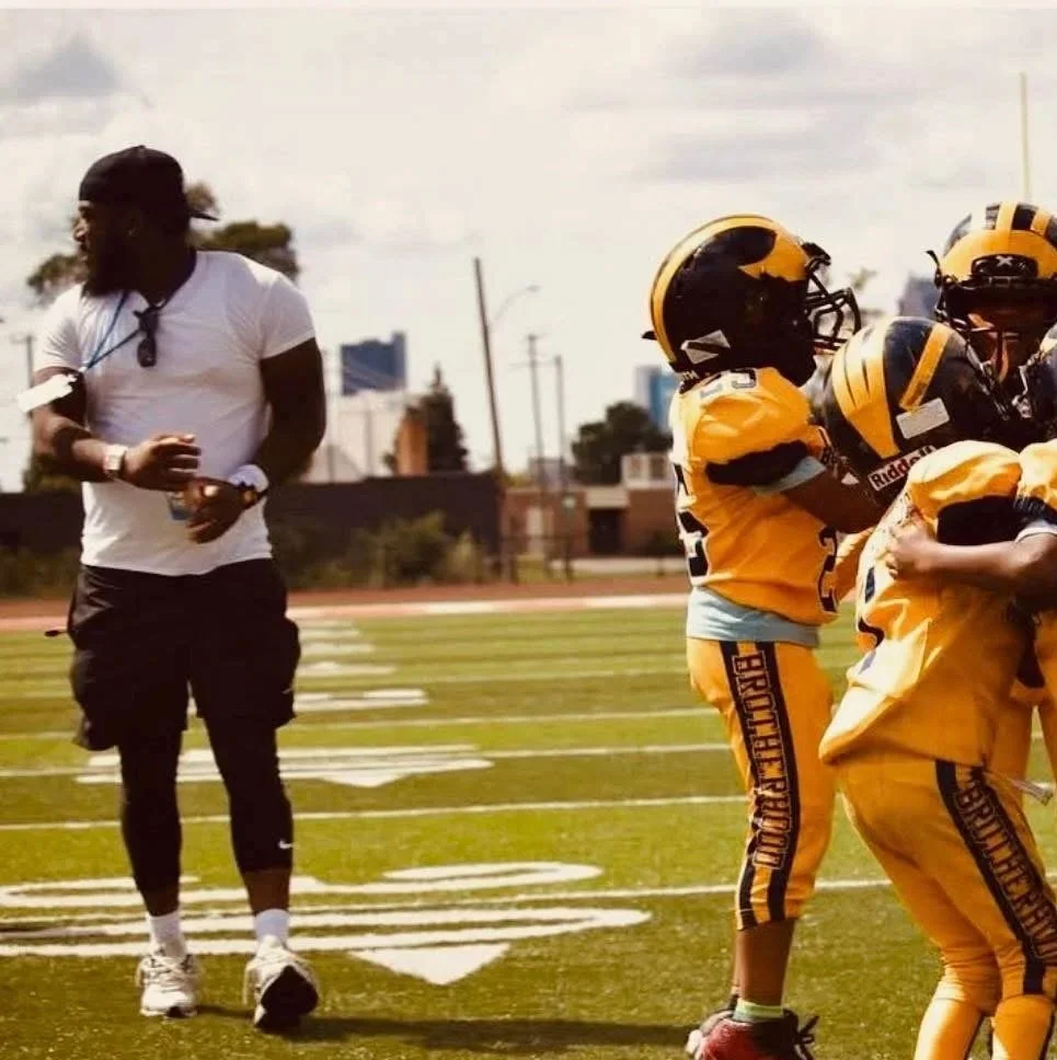 A football coach or staff member standing on a football field near a group of young football players in yellow uniforms and helmets.