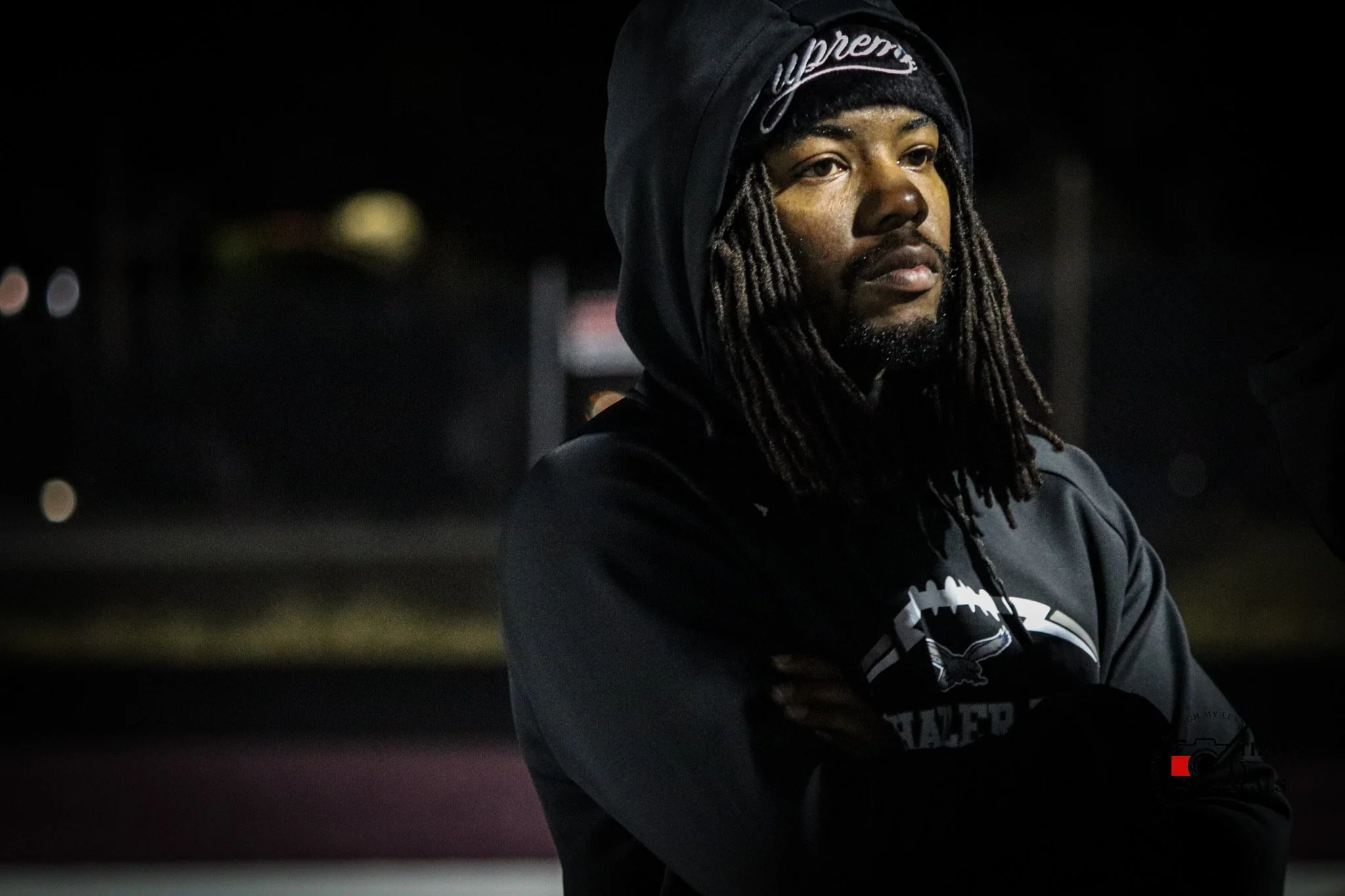 A man with dreadlocks wearing a dark hoodie and a beanie with the Supreme logo, standing with arms crossed in a dark outdoor setting at night.