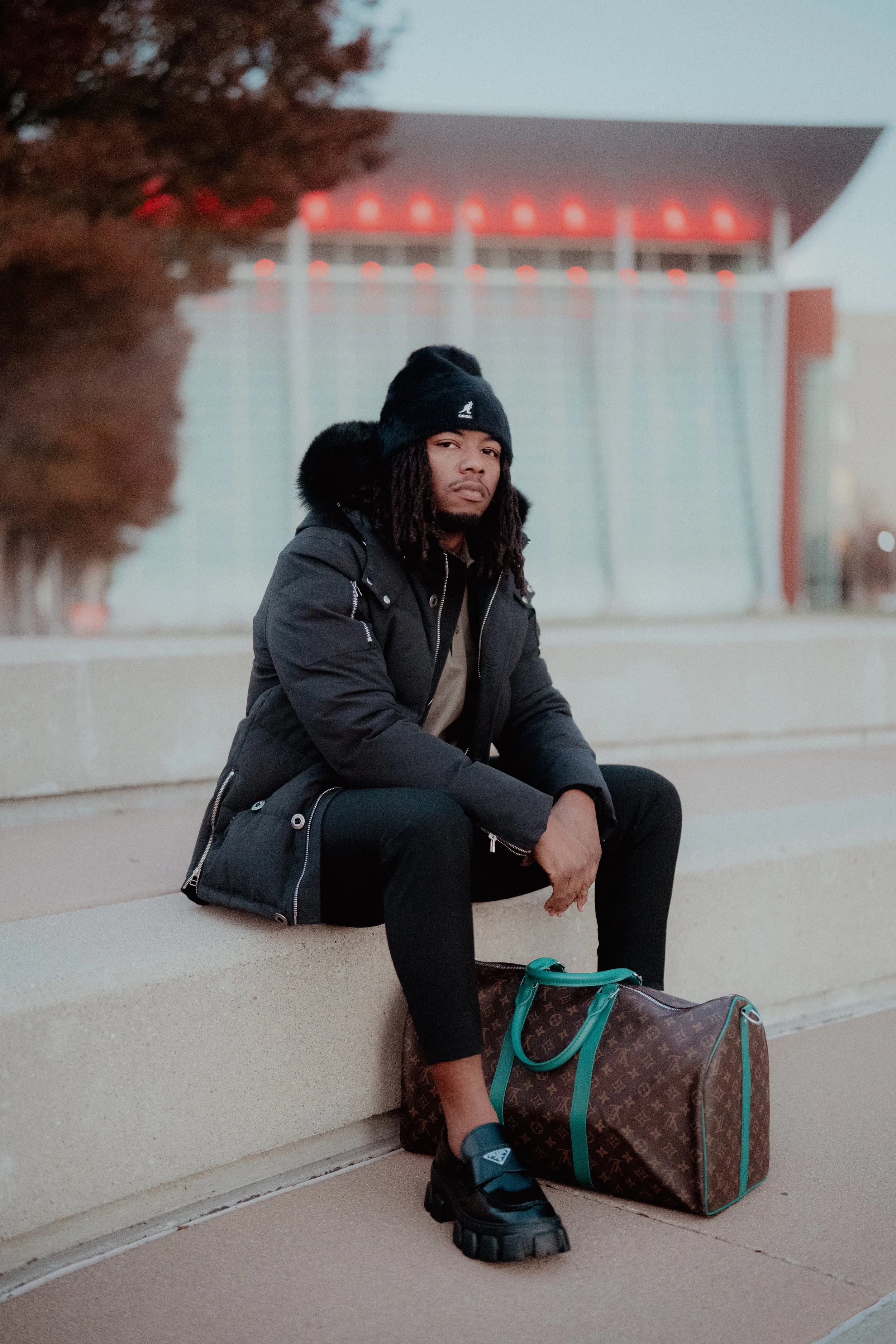 A young man with dreadlocks sits on a concrete bench outdoors, wearing a black winter coat, black beanie, and black shoes, with a Louis Vuitton duffel bag on the ground next to him and a black backpack beside him. A modern building with red lighting is in the background.